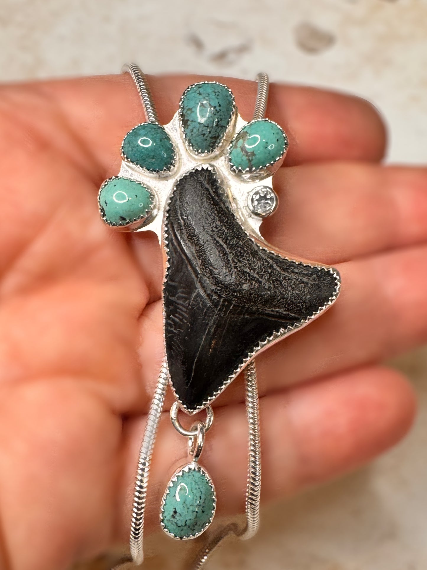 Sterling silver statement, Bolo Necklace with a megalodon fossil tooth, white sapphire and green turquoise held in a hand.