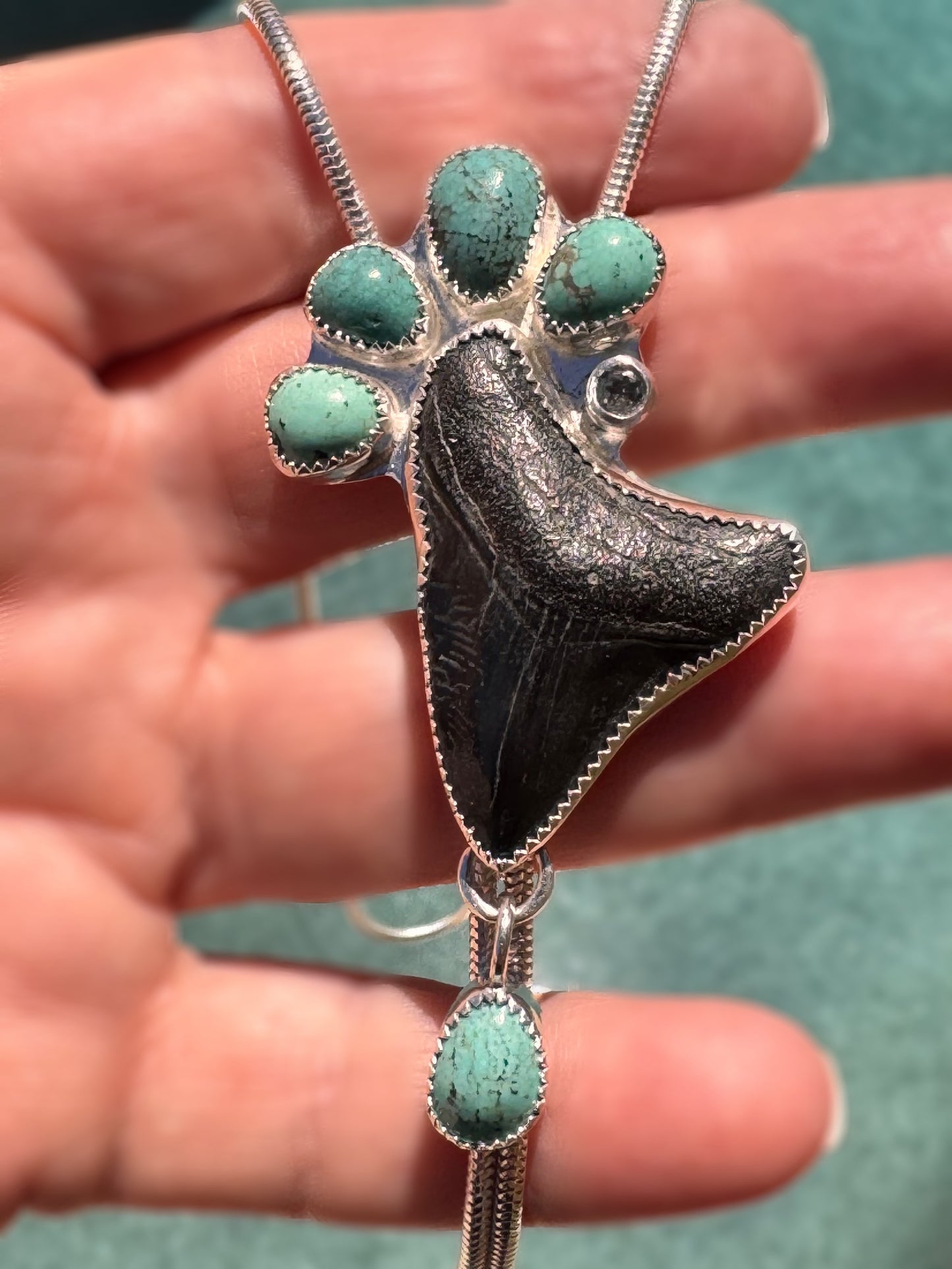 Jewelry pendant with turquoise stones and silver accents held in a hand against a blurred background