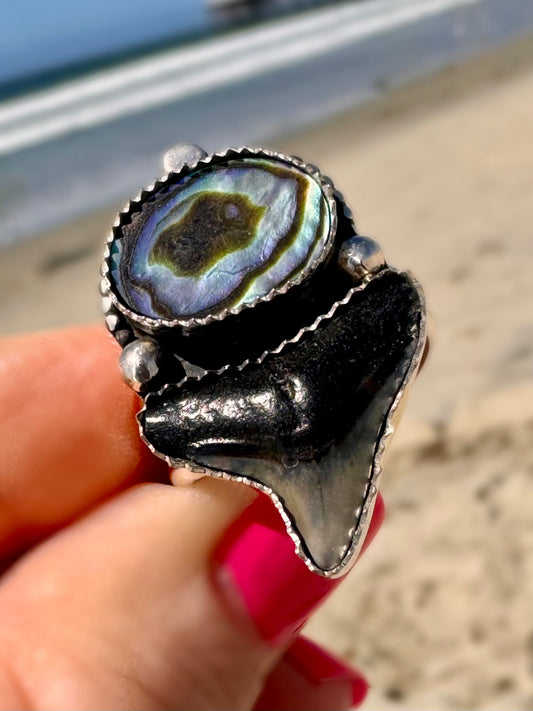 Silver ring with a abalone shell and fossil shark tooth held by a hand on a blurred beach background