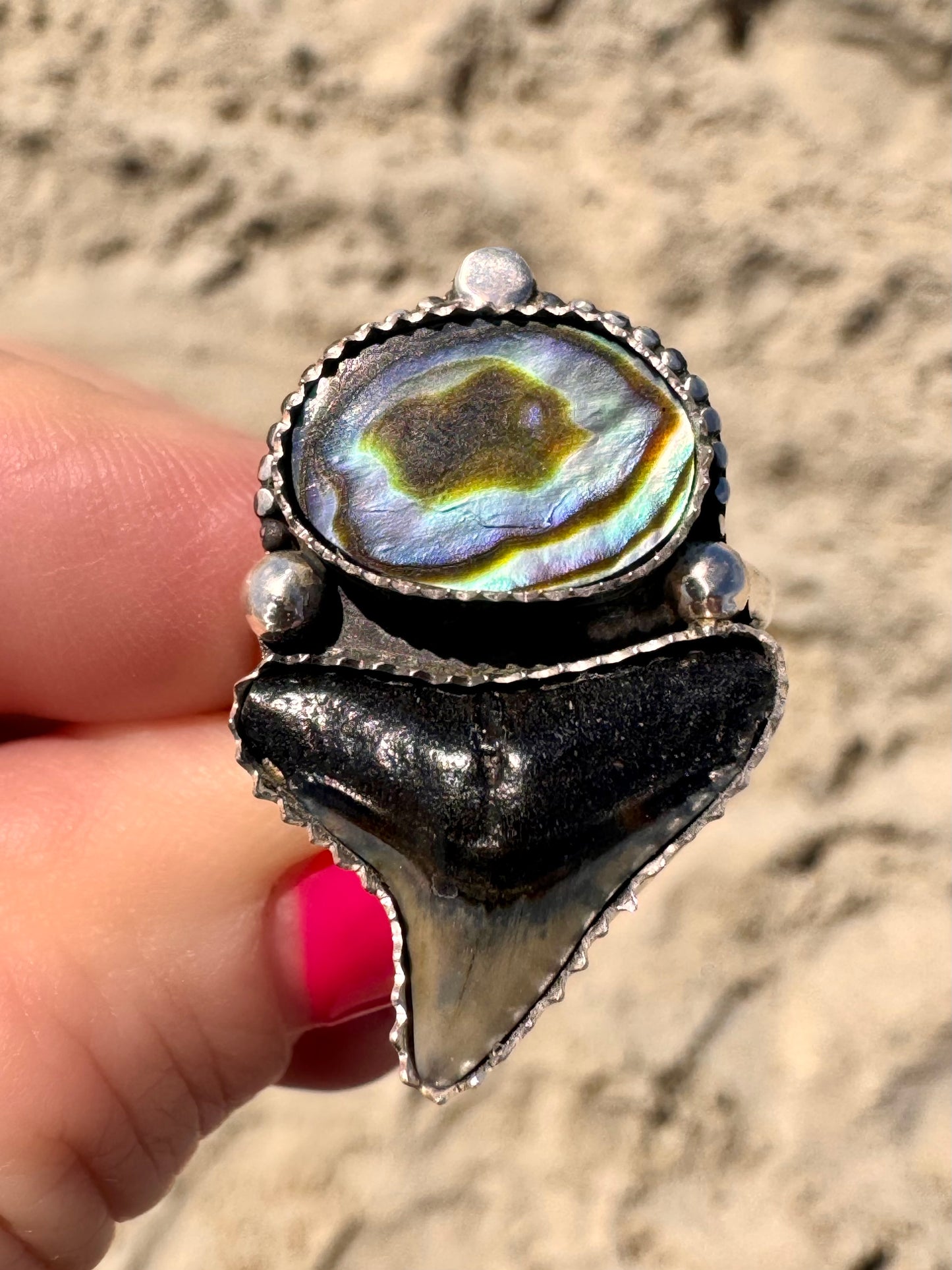 Ring with abalone shell and shark tooth held by a hand on a sandy background