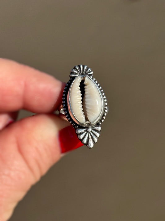 Silver ring with a central shell design held by a hand against a neutral background