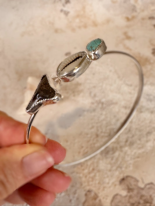 Silver bracelet with shell, turquoise and shark tooth design held by a hand against a beige background