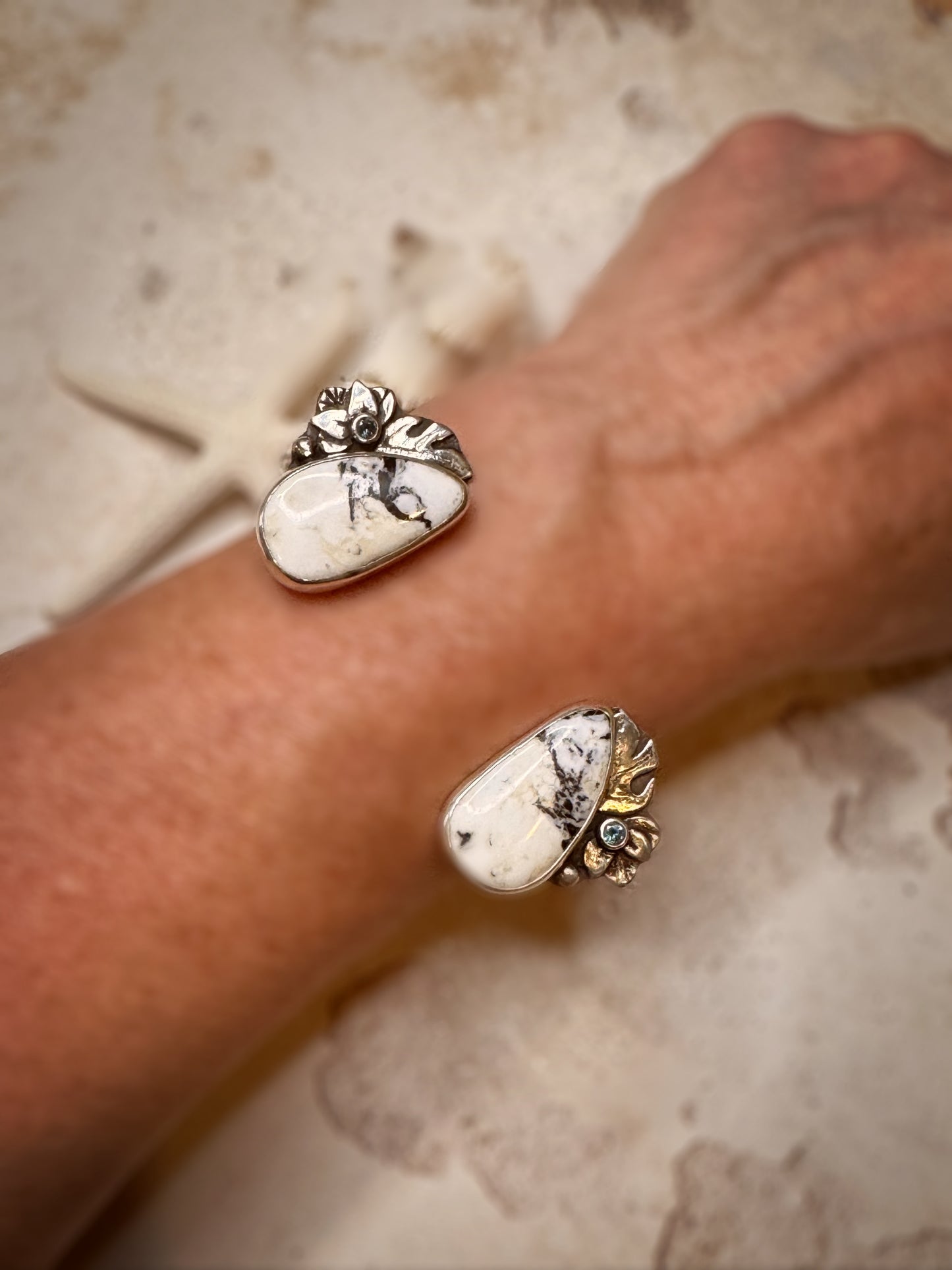 Close-up of a wrist wearing a silver, white buffalo, Montana sapphire and monstera leaf cuff on a textured surface.