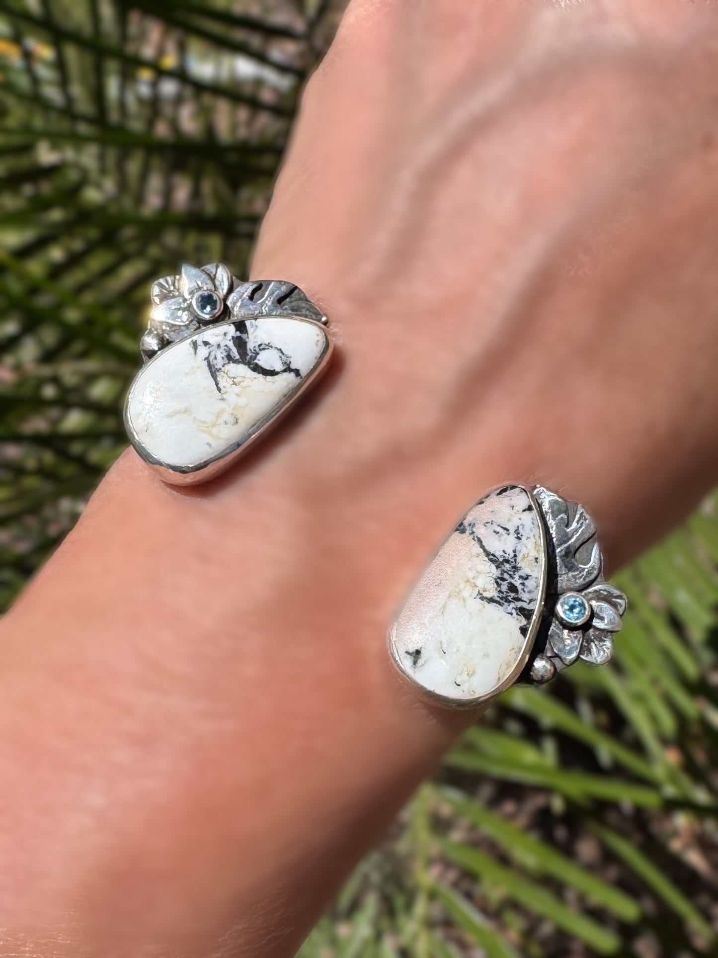 Close-up of a wrist wearing a silver, white buffalo, Montana sapphire and monstera leaf cuff against a palm tree.