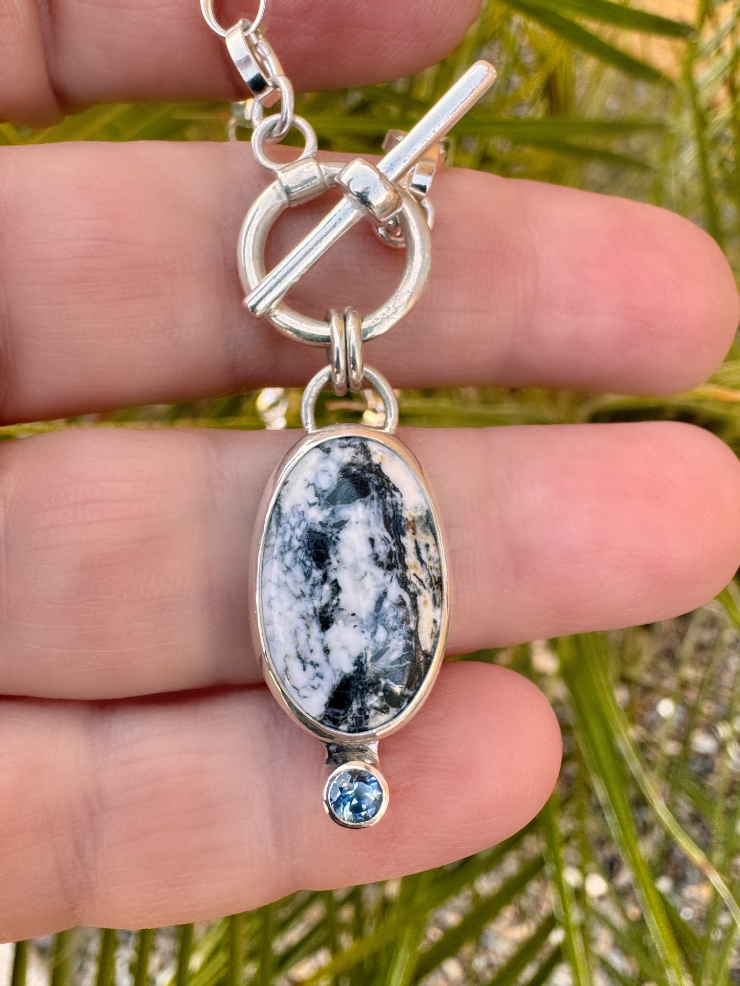 Close-up photo of sterling silver necklace with oval White Buffalo stone and teal Montana sapphire accent on a 16-inch toggle chain.