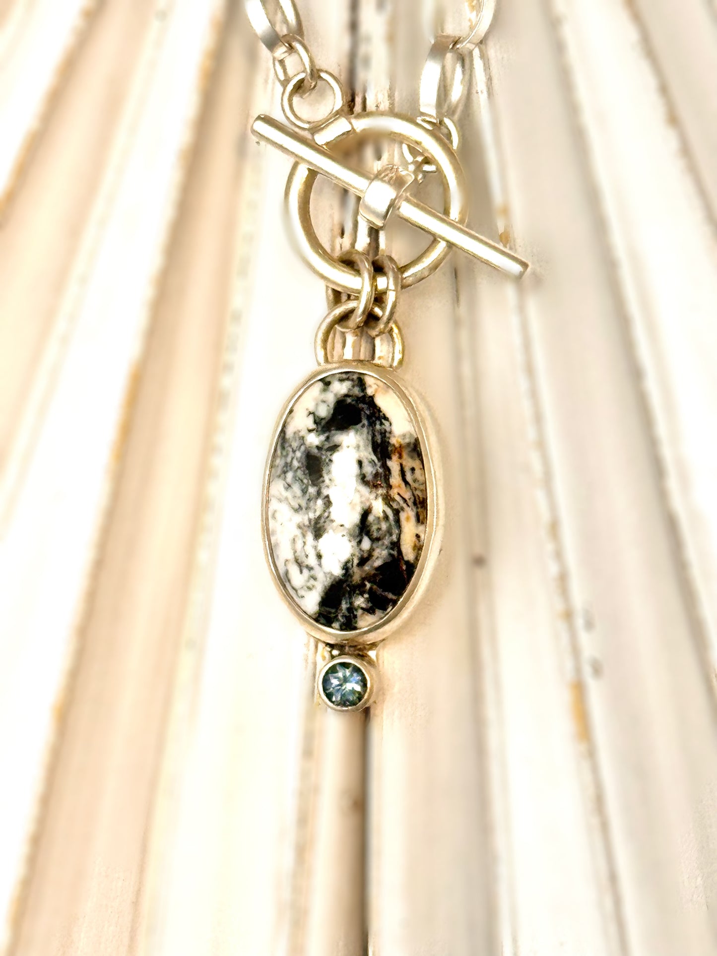 Close-up photo of sterling silver necklace with oval White Buffalo stone and teal Montana sapphire accent on a 16-inch toggle chain.