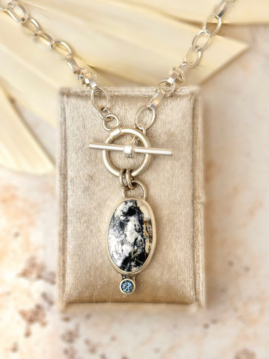 Close-up photo of sterling silver necklace with oval White Buffalo stone and teal Montana sapphire accent on a 16-inch toggle chain.