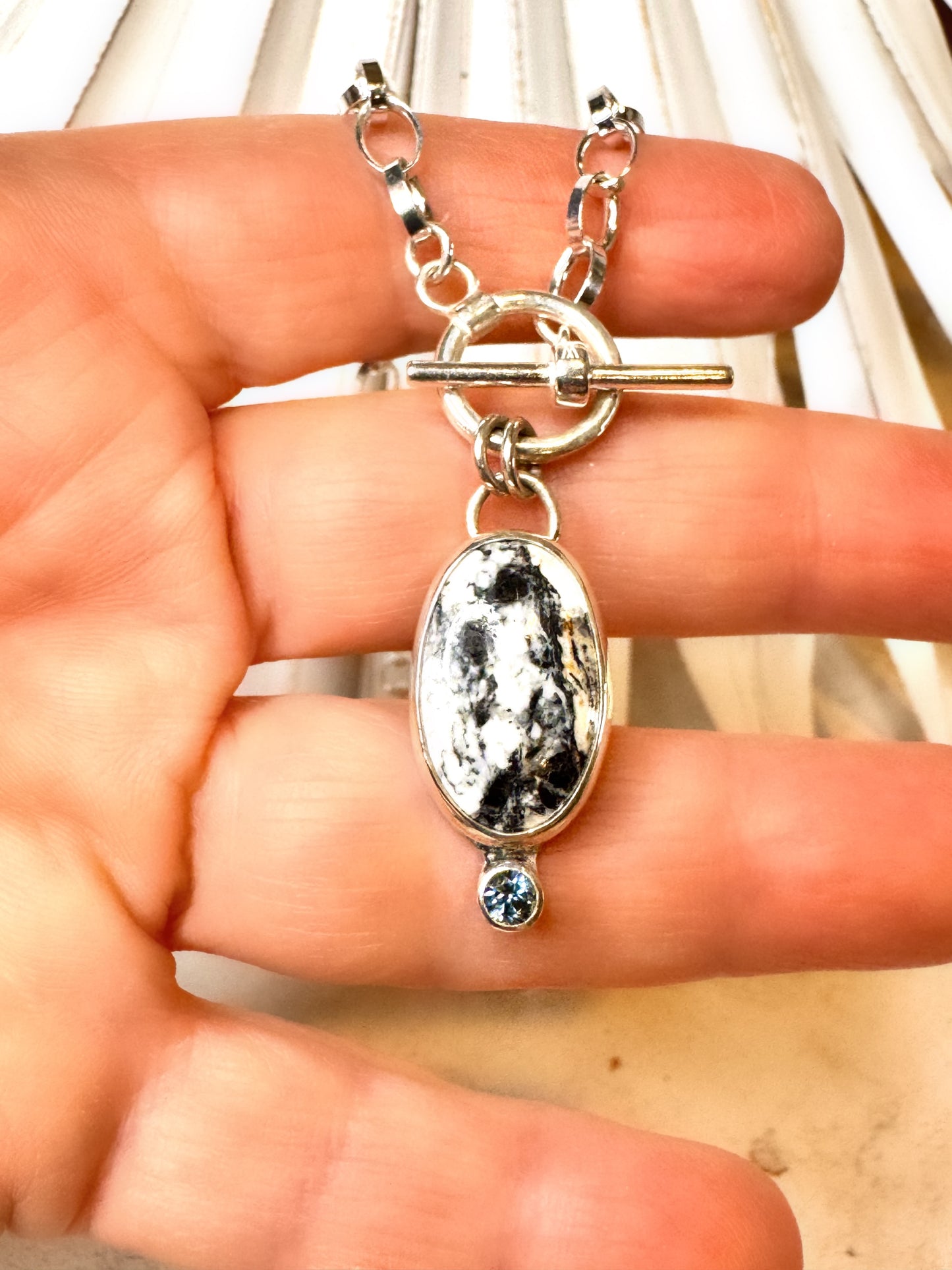 Close-up photo of sterling silver necklace with oval White Buffalo stone and teal Montana sapphire accent on a 16-inch toggle chain.