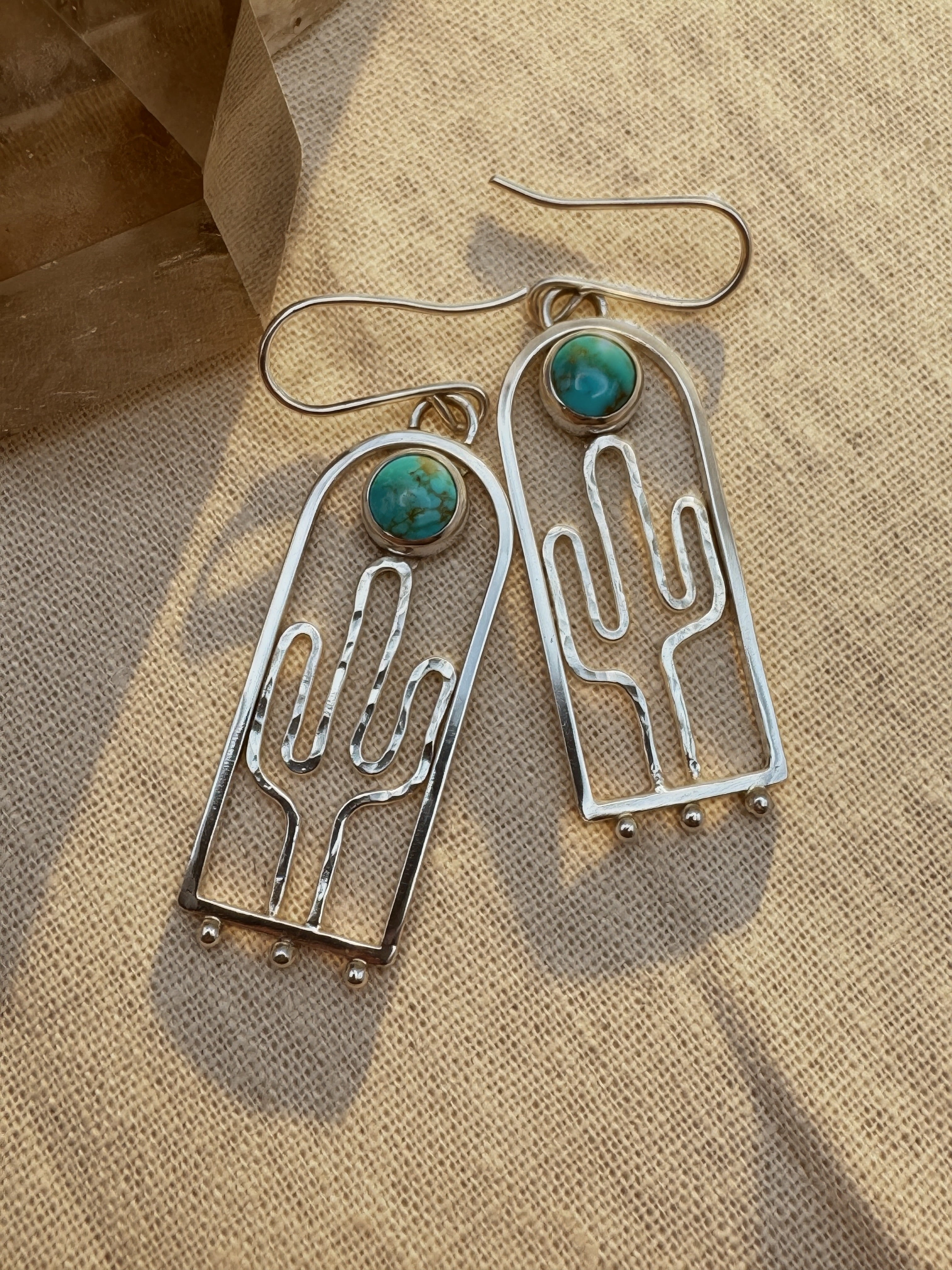 Hand-hammered sterling silver saguaro earrings with Blue Moon turquoise cabochons, approximately 2.5 inches, on a warm neutral background