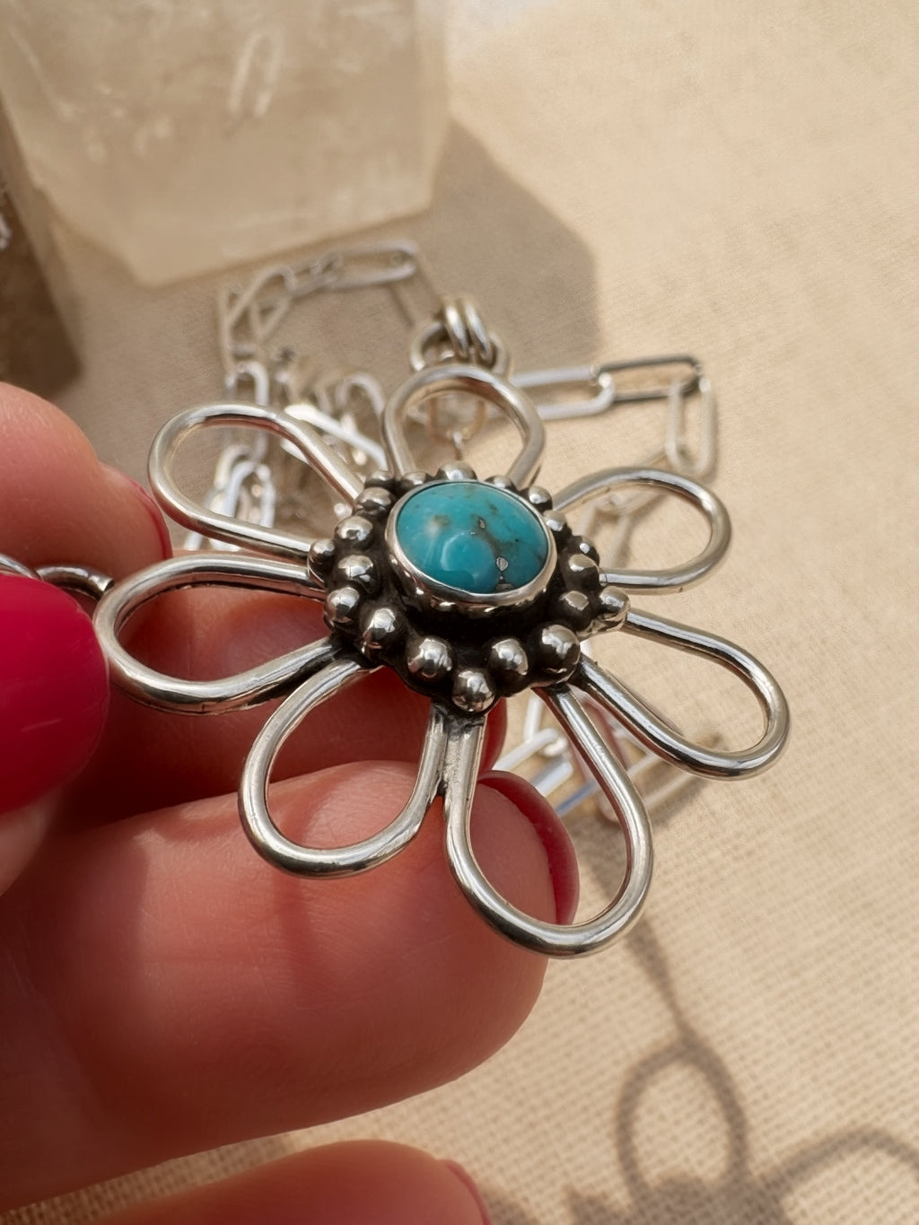 Silver flower-shaped pendant with a sleeping beauty turquoise center held by a hand against a beige background