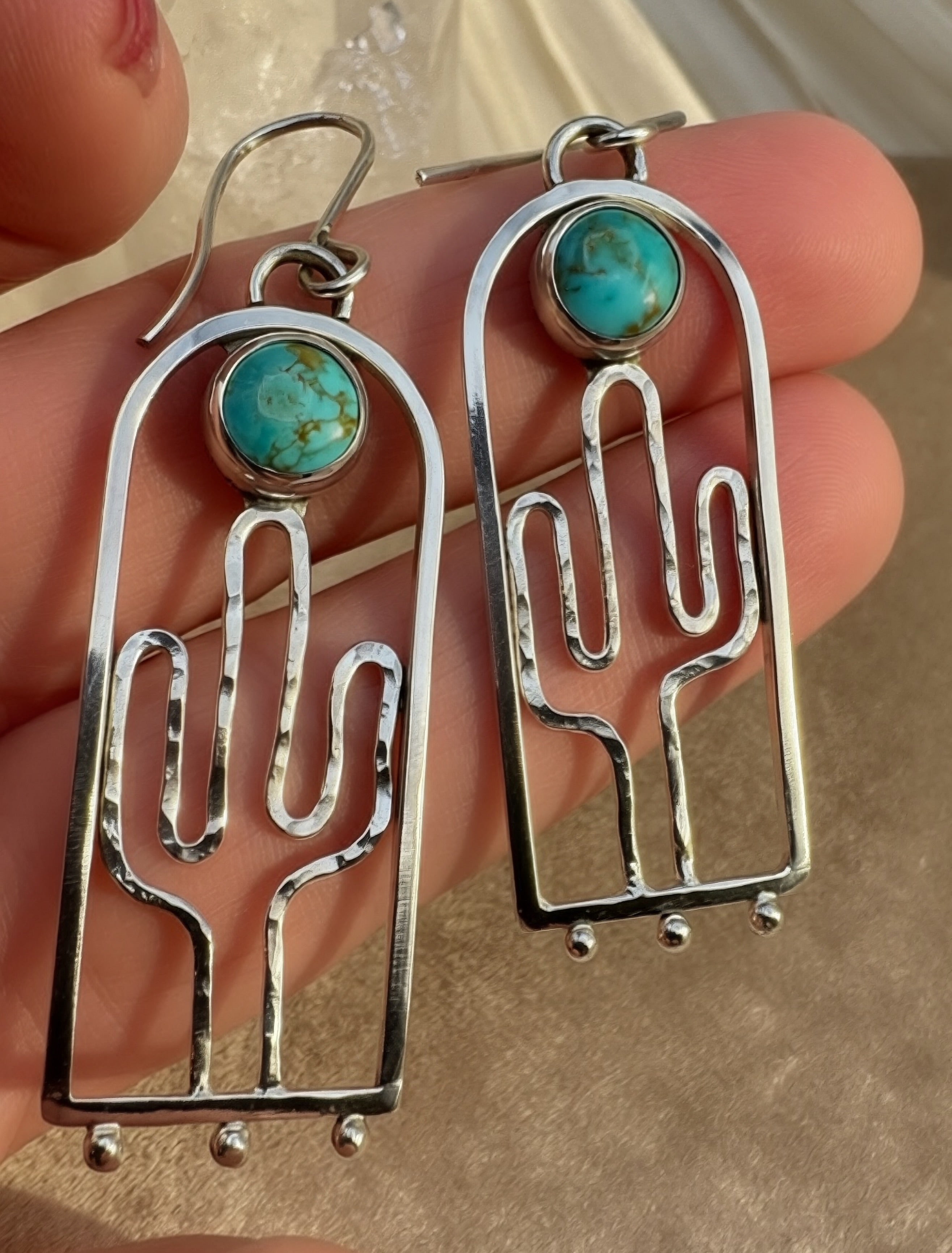 Hand-hammered sterling silver saguaro earrings with Blue Moon turquoise cabochons, approximately 2.5 inches, in hand on a warm neutral background