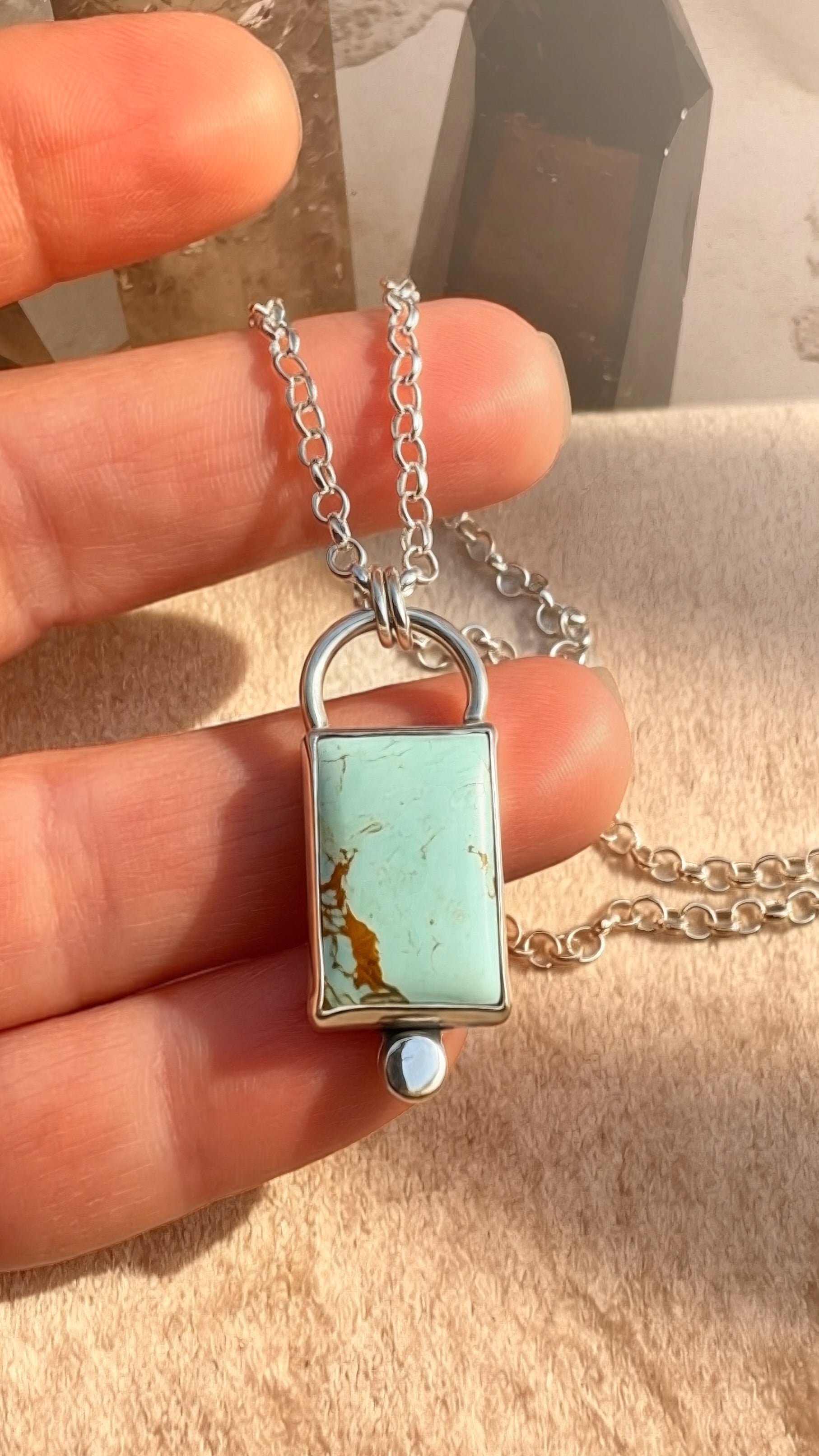 Silver lock-shaped pendant with turquoise stone held between fingers on a beige surface