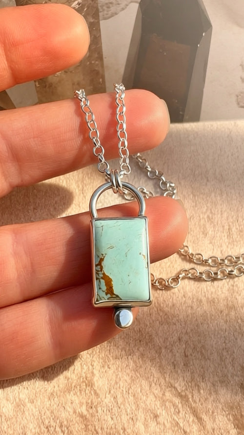 Silver lock-shaped pendant with turquoise stone held between fingers on a beige surface