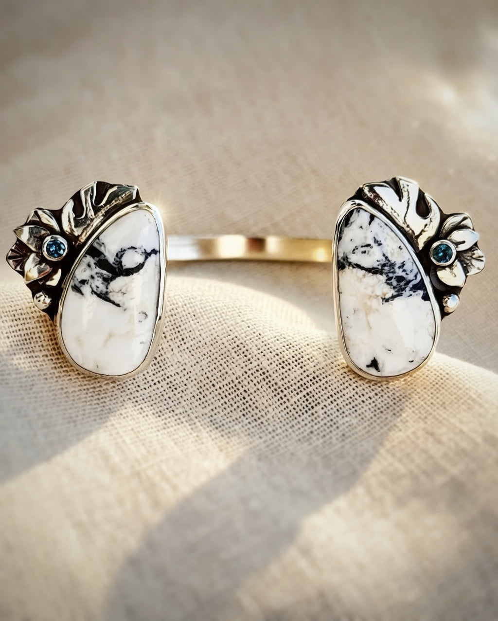 A silver  cuff bracelet with two large white and black marbled White Buffalo stone cabochons, each accented with a small round Montana sapphire with Monstera and flower silver detailing, displayed on a textured beige fabric.