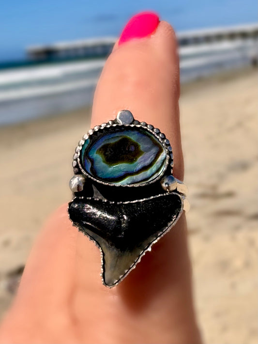 Close-up of a hand wearing a large ring with a unique design with abalone shell and a fossil shark tooth, set against a beach background.