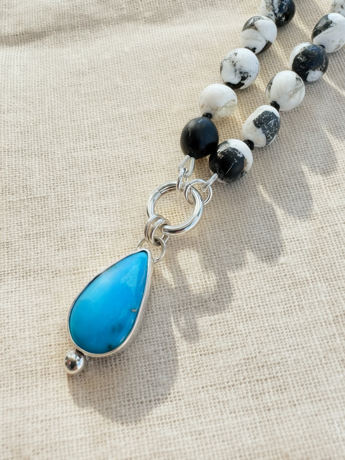 Blue Meridian Arizona turquoise pendant and charm keeper beside black-and-white composite beads on beige fabric.