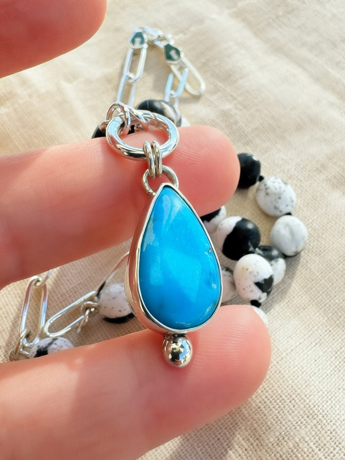 Blue Meridian turquoise pendant held between fingers, showing its teardrop shape and silver droplet detail.
