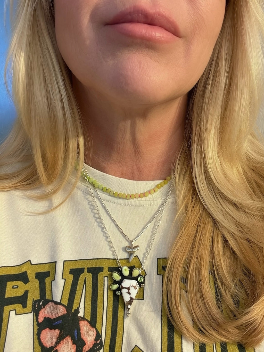 Close-up of a person wearing layered necklaces, including a silver chain with a green, brown and white pendant and a yellow-green beaded necklace, over a cream graphic t-shirt.
