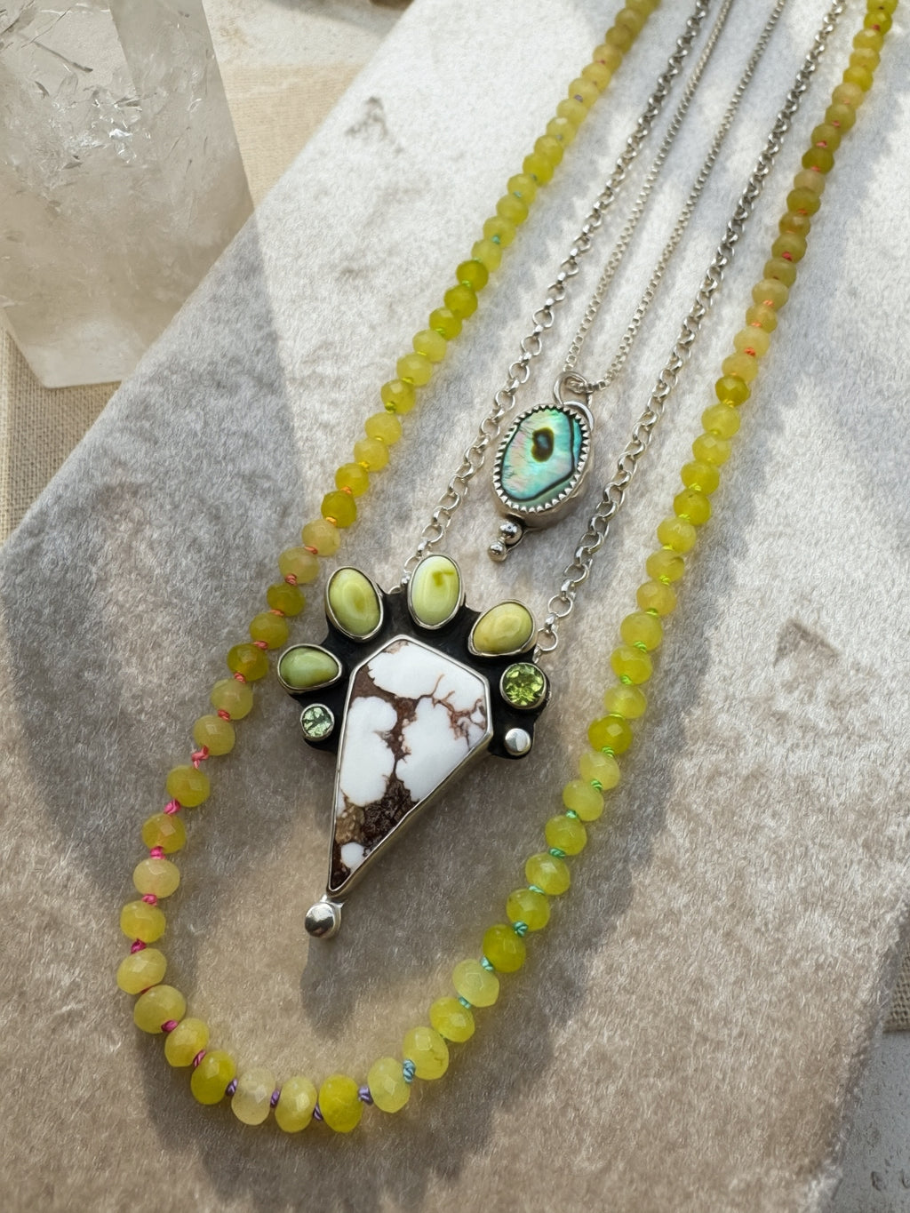 A layered necklace set displayed on a textured neutral surface. The set features a long strand of yellow-green beads, a silver chain with a triangular white and brown stone pendant surrounded by small green variscite and peridot stones, and a smaller oval abalone pendant on a silver chain.