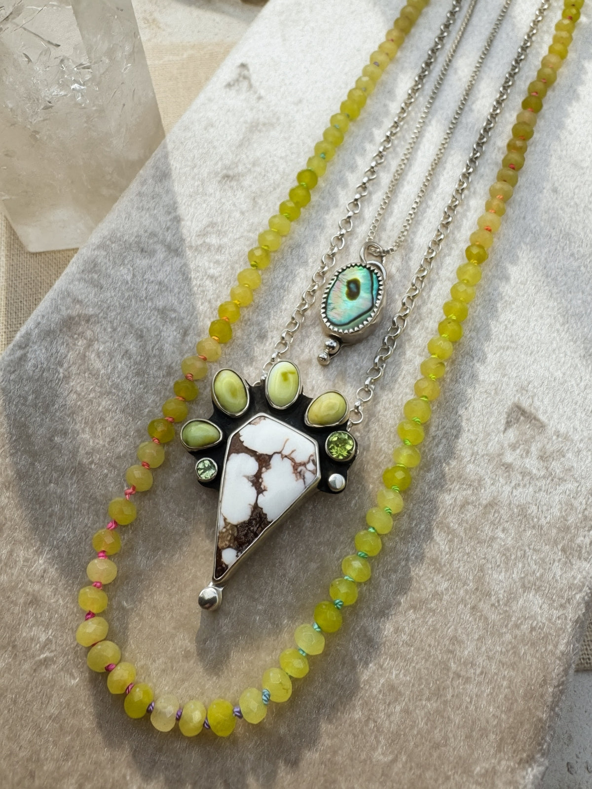 A layered necklace set displayed on a textured neutral surface. The set features a long strand of yellow-green beads, a silver chain with a triangular white and brown stone pendant surrounded by small green variscite and peridot stones, and a smaller oval abalone pendant on a silver chain.
