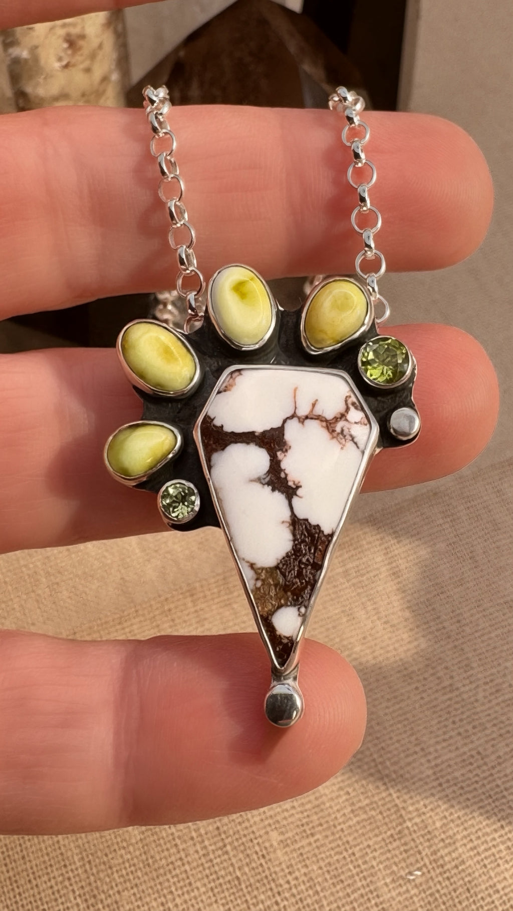 A close-up of a hand holding a silver pendant necklace featuring a triangular white and brown stone centerpiece, surrounded by small oval yellow-green stones and clear round stones on a silver chain.
