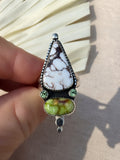 A hand holding a silver ring featuring a large white and brown marbled stone, a green stone, and small green accent stones, set against a neutral fabric background.