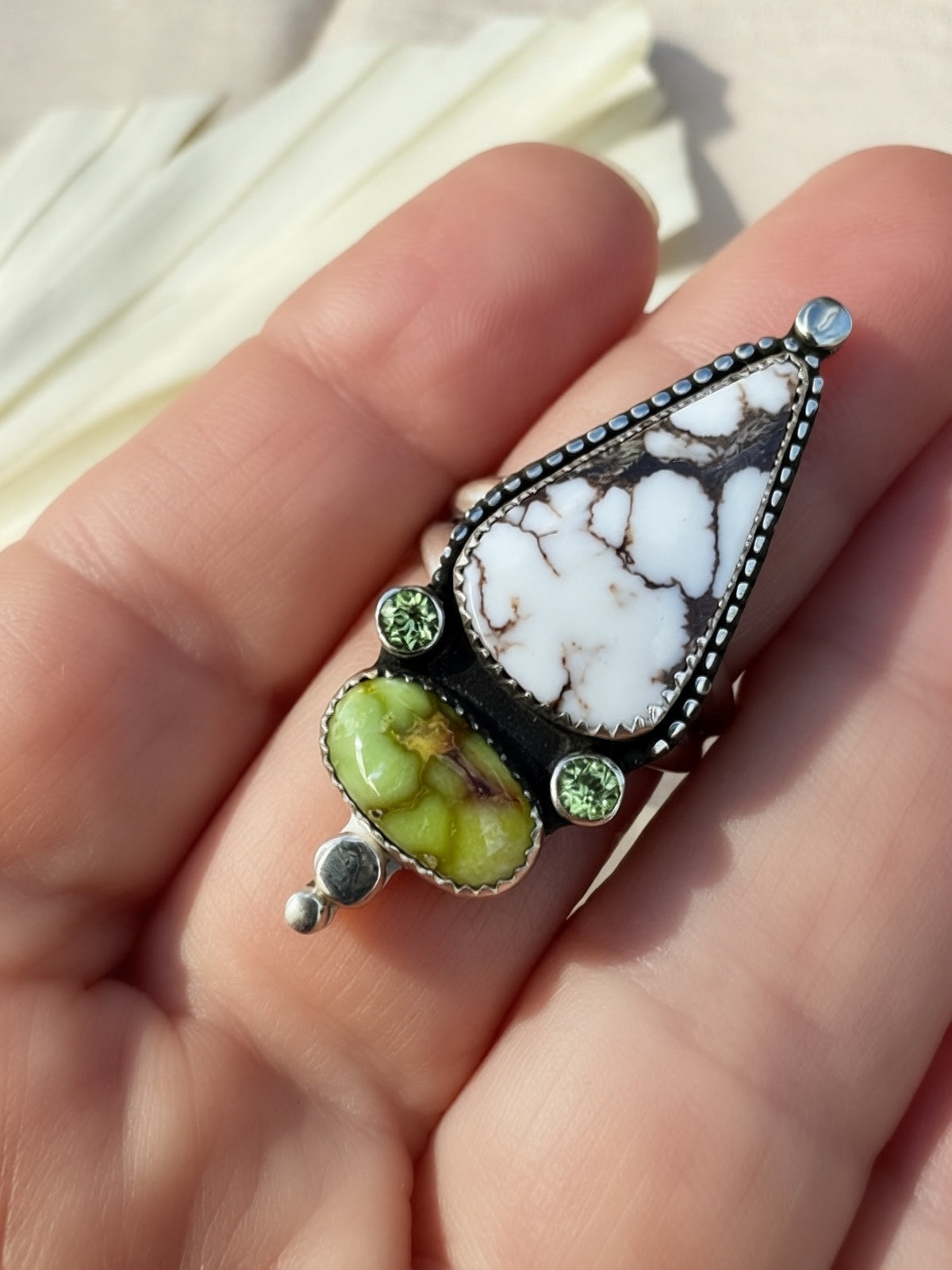 A close-up of a hand holding a silver statement ring featuring a large white marbled stone, a green variscite stone, and small green peridot accent gemstones.