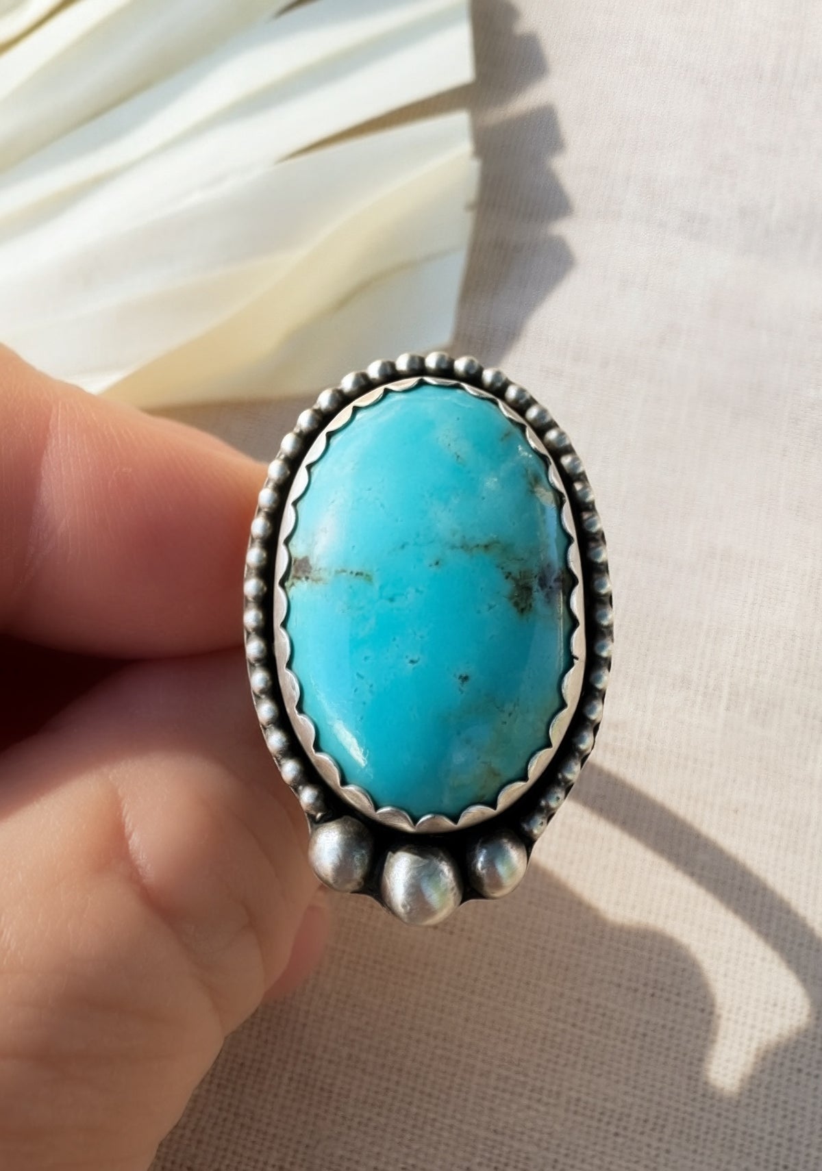 A hand holding a sterling silver ring featuring a large oval turquoise cabochon stone with natural matrix inclusions, set against a neutral beige fabric background.