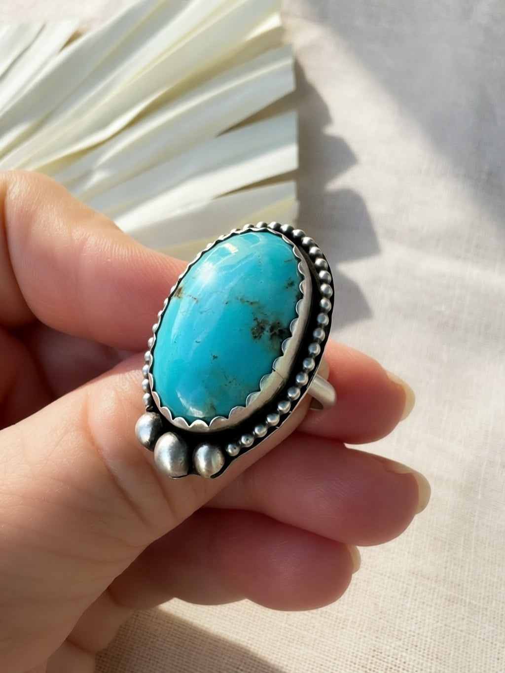 A hand holding a sterling silver ring with a large oval turquoise cabochon, set against a neutral beige fabric background with a decorative white fan in the upper left.