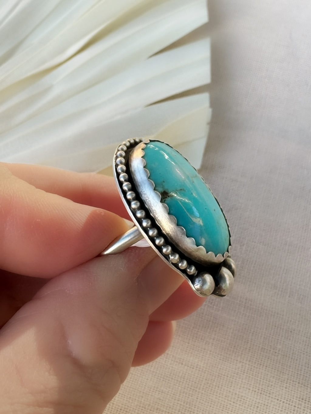 A sterling silver ring with an oval turquoise cabochon stone held by a hand against a neutral beige fabric background.