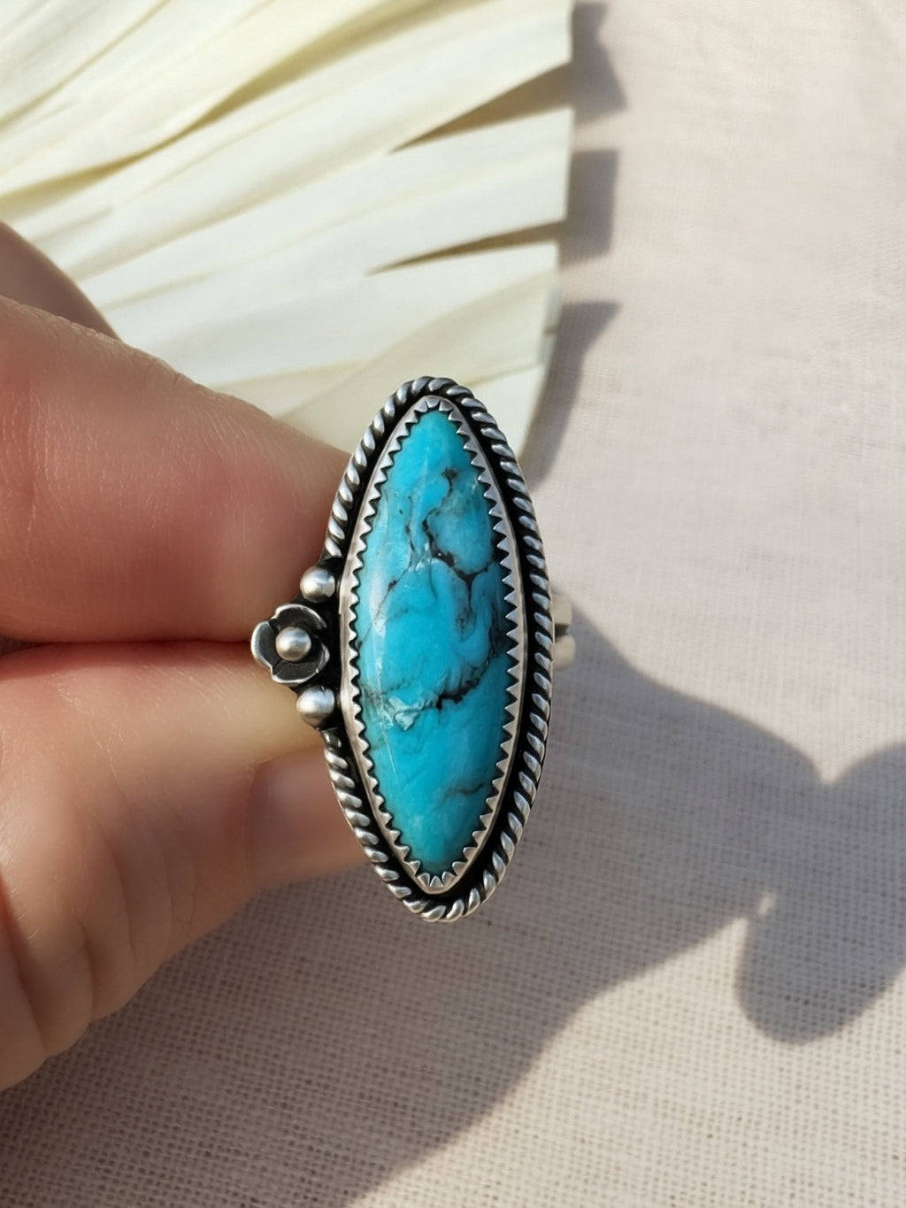A hand holding a silver ring with a large oval turquoise stone, photographed against a light fabric background with a decorative fan behind it.