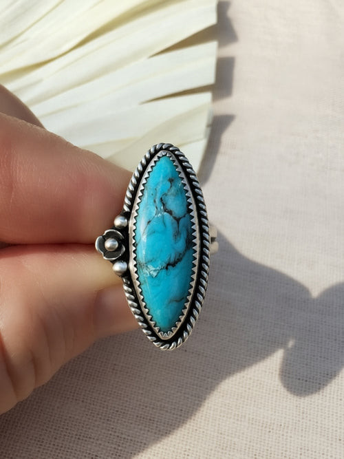 A hand holding a silver ring with a large oval turquoise stone, photographed against a light fabric background with a decorative fan behind it.