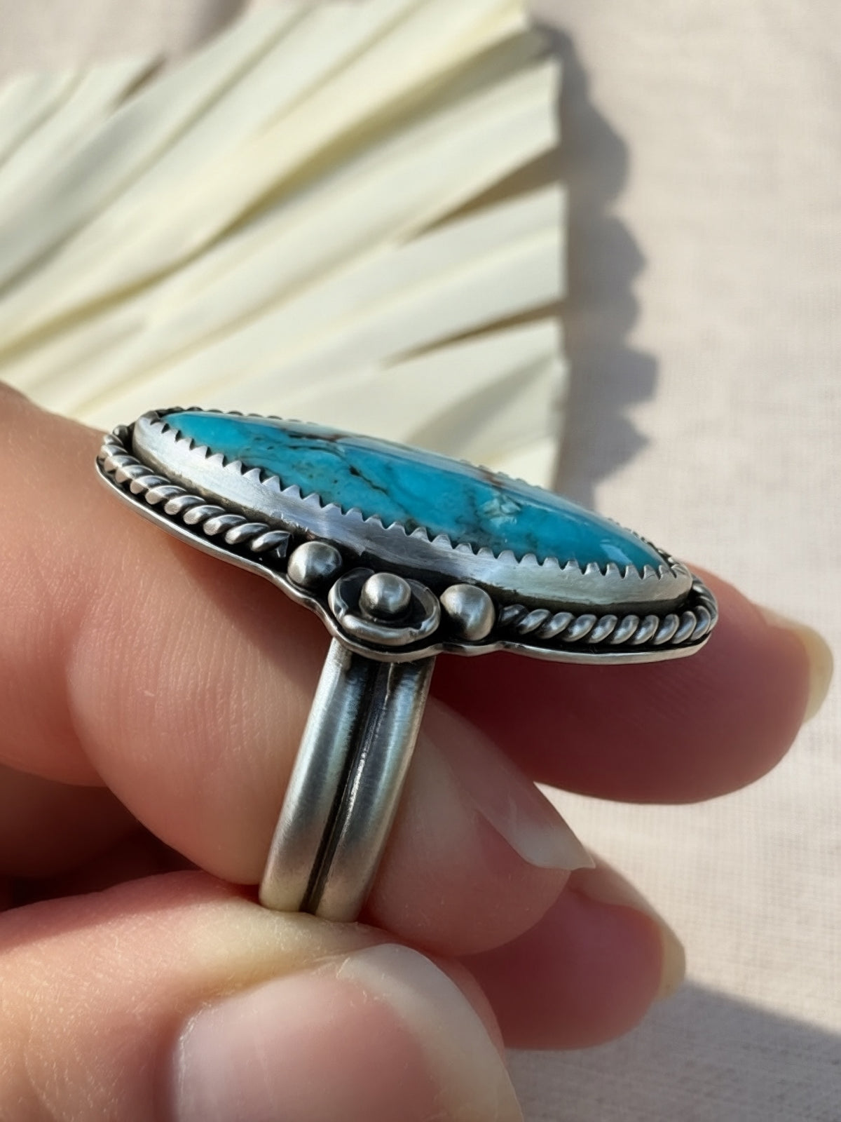 A sterling silver ring with a large oval turquoise stone, held on a person's finger against a light beige background with a decorative fan blurred in the background.
