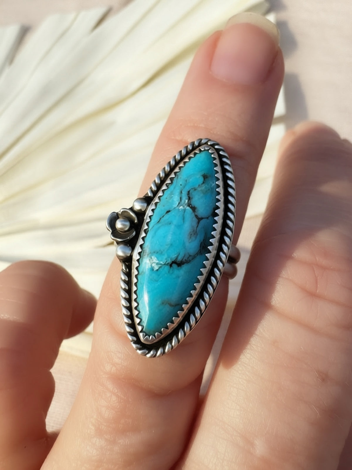 A close-up of a hand wearing a silver ring with a large oval turquoise stone. The ring features a textured, braided silver border and a small decorative flower and silver beads on the left side, set against a soft-focus background of light-colored dried leaves.