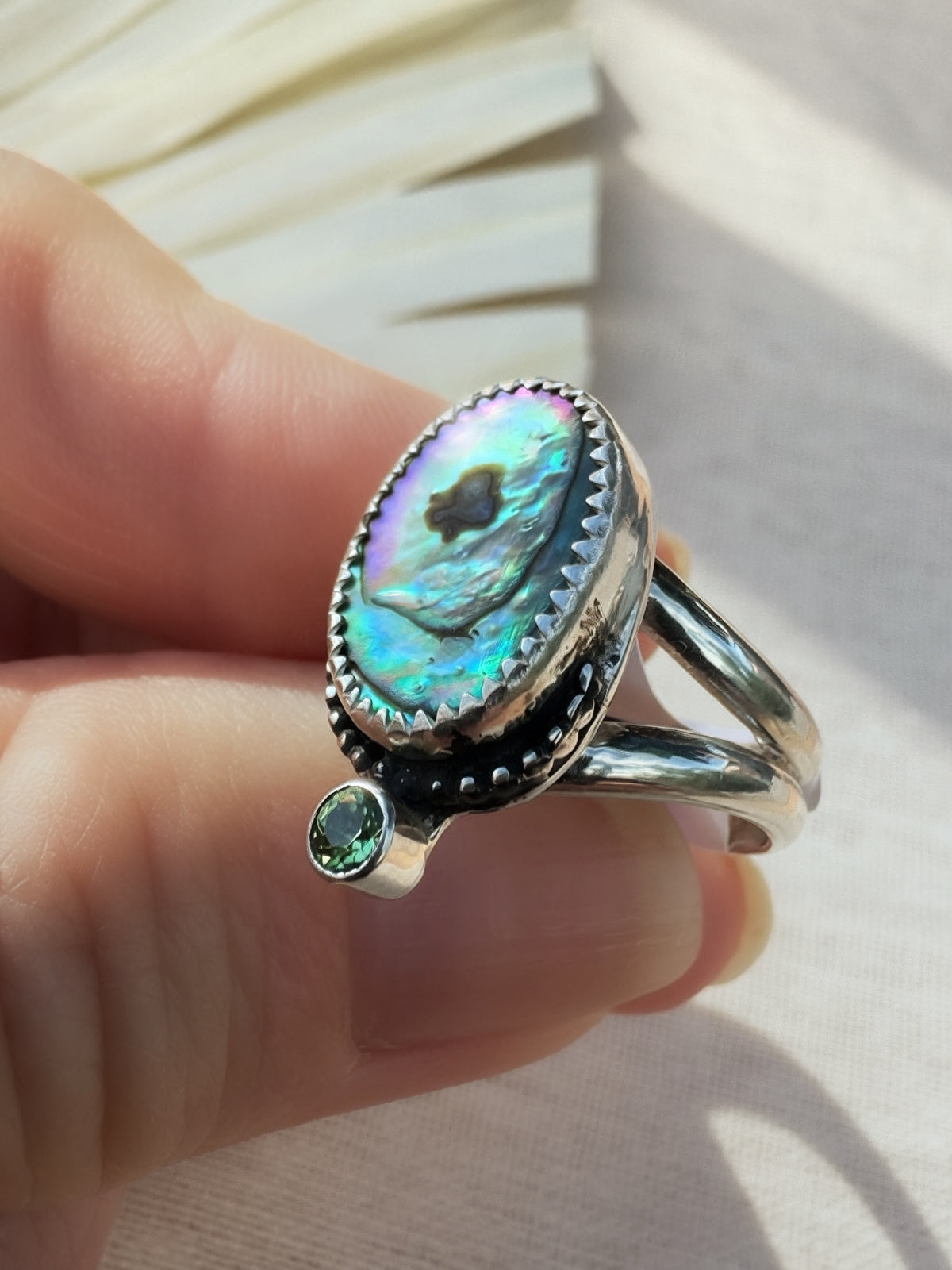 A sterling silver ring with an oval iridescent abalone shell centerpiece and a green peridot gemstone accent, shown being held against a light fabric background.