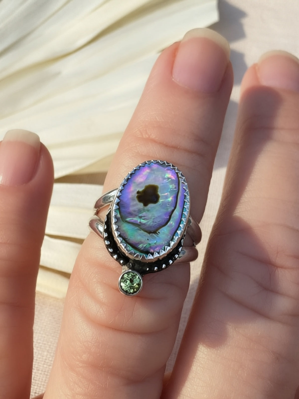 A silver ring with an oval iridescent abalone shell centerpiece and a small peridot gemstone accent, shown worn on a person's finger.
