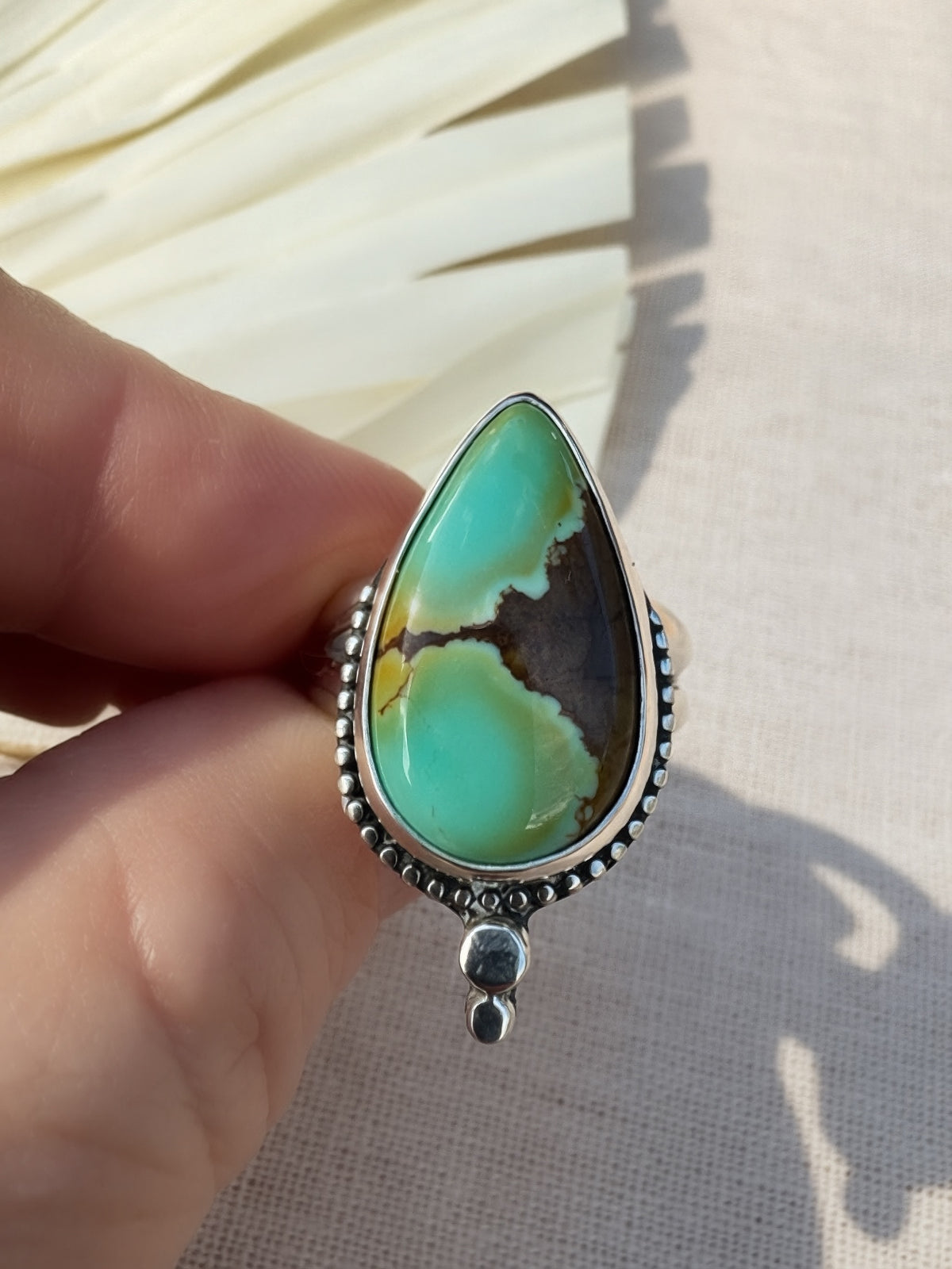 A hand holding a silver ring featuring a large teardrop-shaped turquoise stone with natural brown and black veining, set against a neutral fabric background.