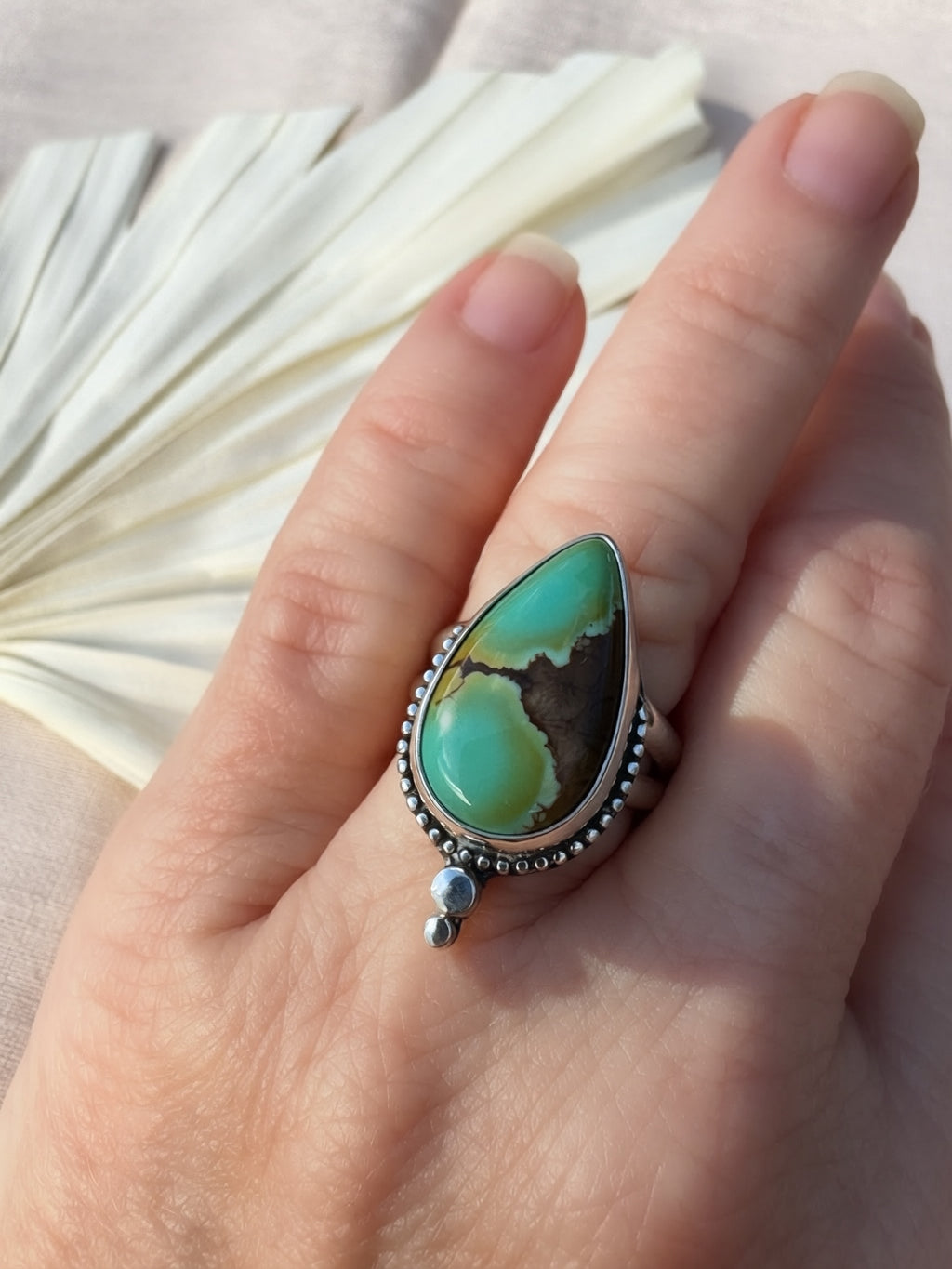 A close-up of a hand wearing a silver ring with a large teardrop-shaped turquoise stone featuring green, brown, and black marbling, with silver bead accents.