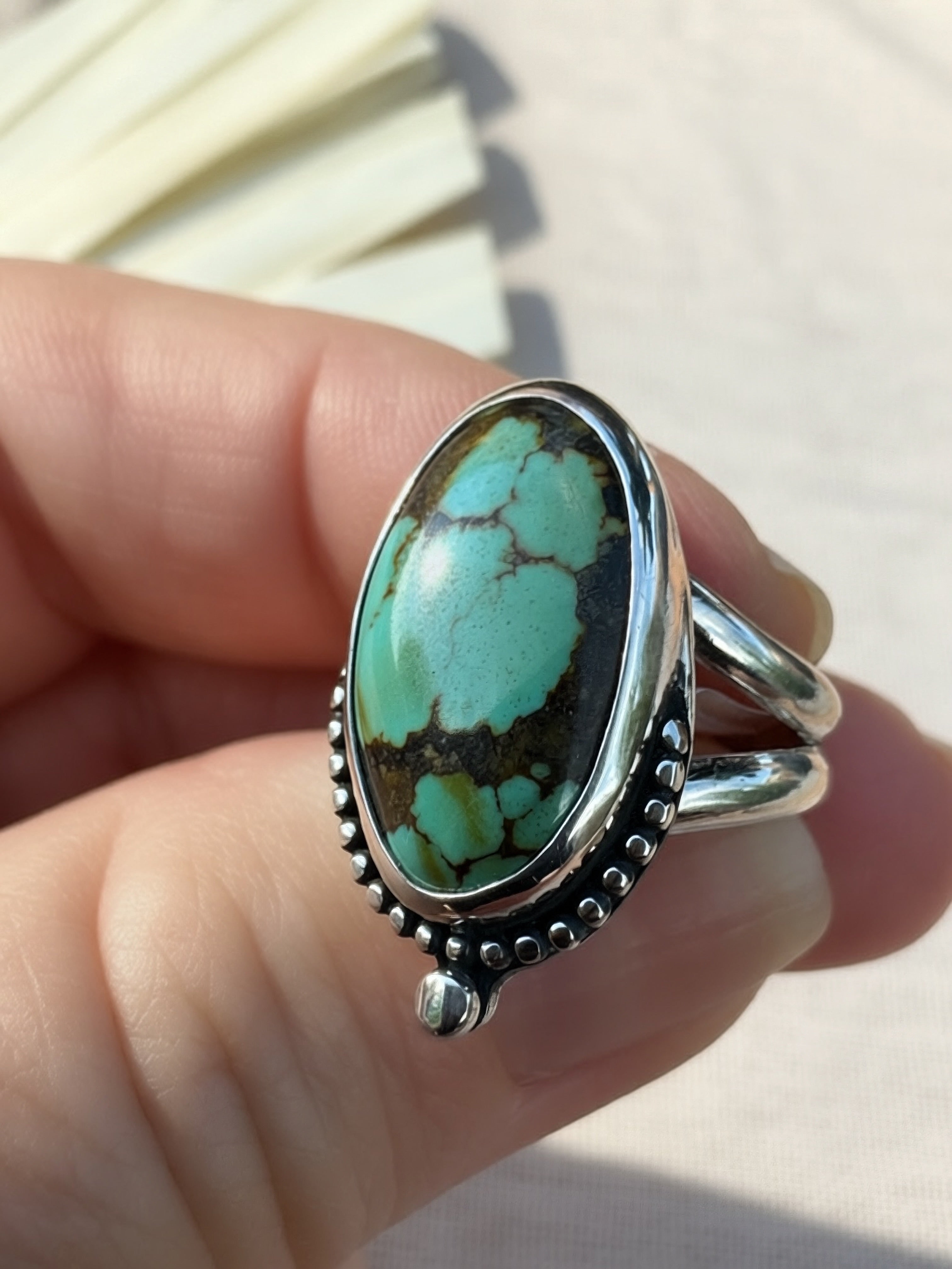 A sterling silver ring with a large oval turquoise stone held on a person's fingers. The ring features a bezel-set oval turquoise cabochon with natural matrix veining, surrounded by a beaded silver border and a split shank band.