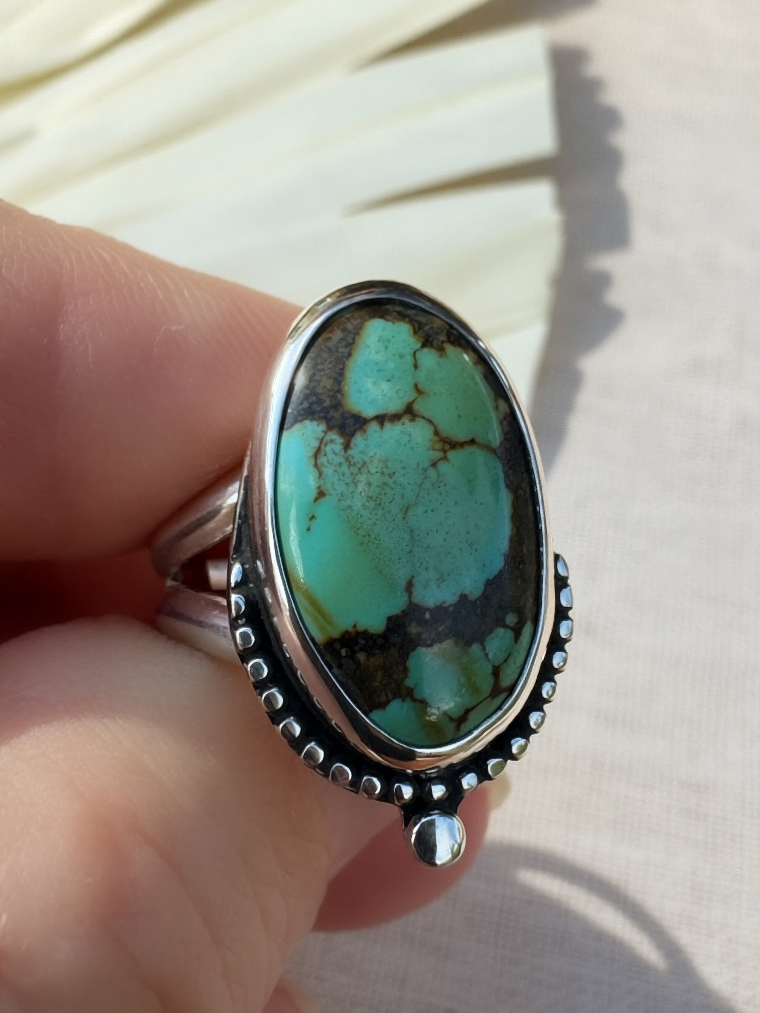 A sterling silver ring with a large oval turquoise stone held by a hand. The ring features a bezel-set turquoise cabochon with natural matrix veining, surrounded by a beaded silver border and a silver bead.