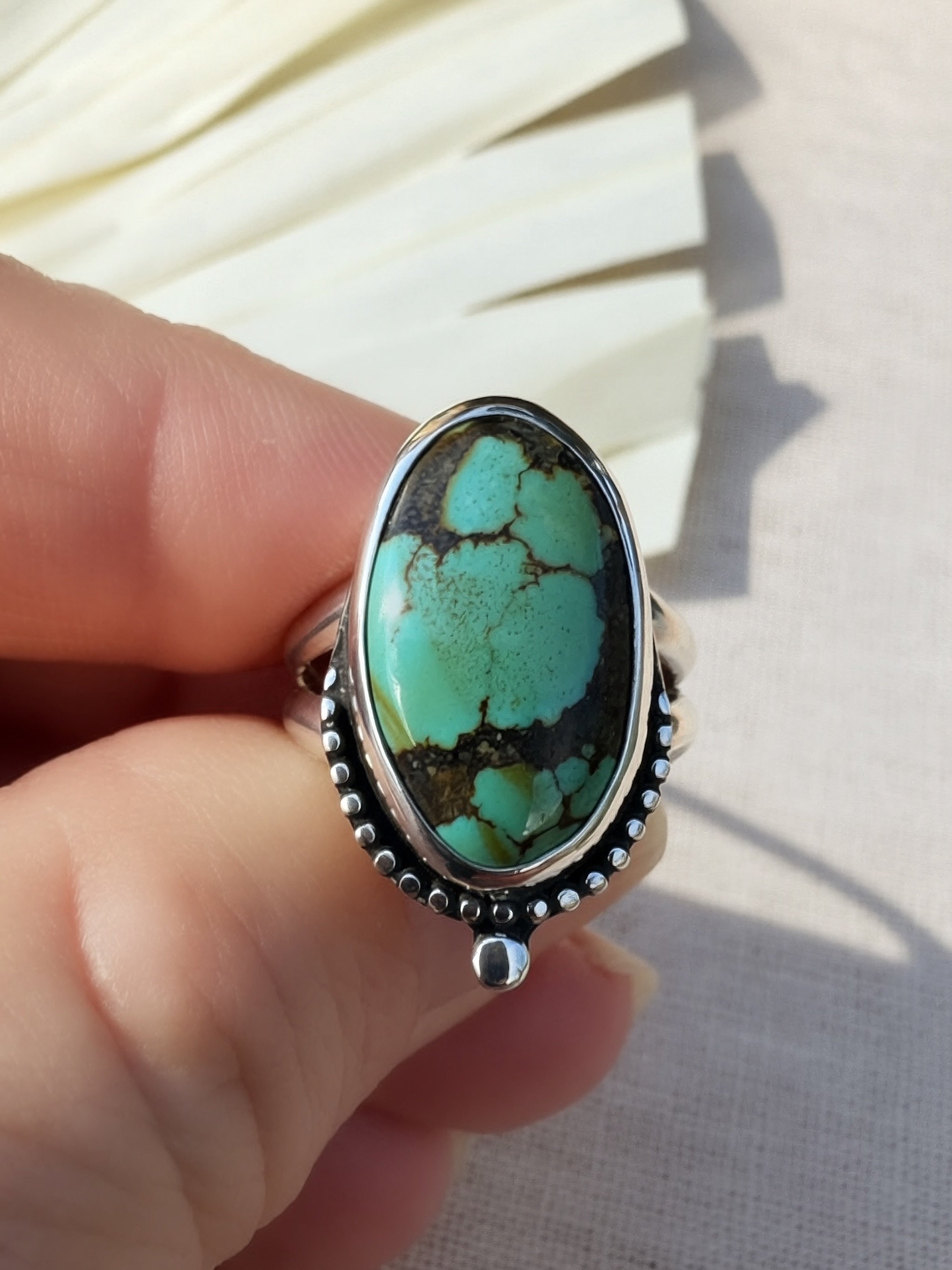 A sterling silver ring with a large oval turquoise stone held by a hand against a neutral beige background.
