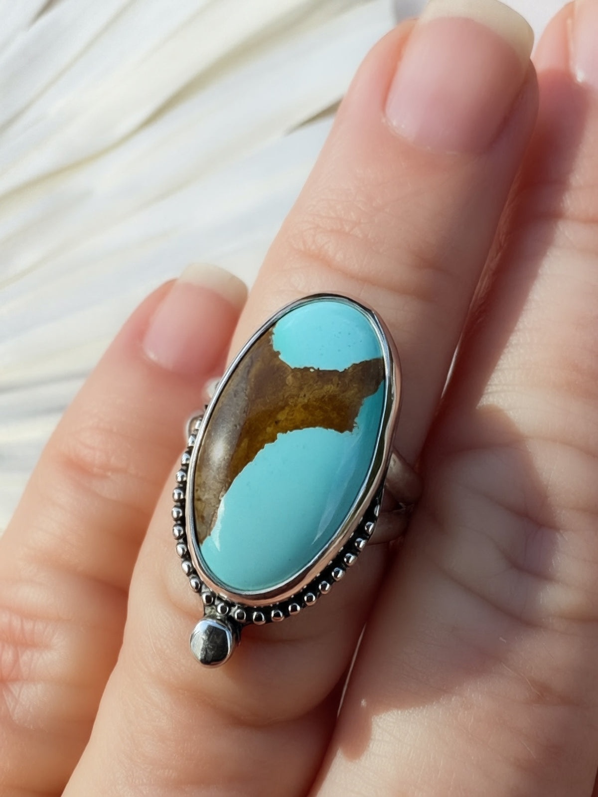 A close-up of a hand wearing a silver ring with an pale blue oval turquoise with brown matrix.