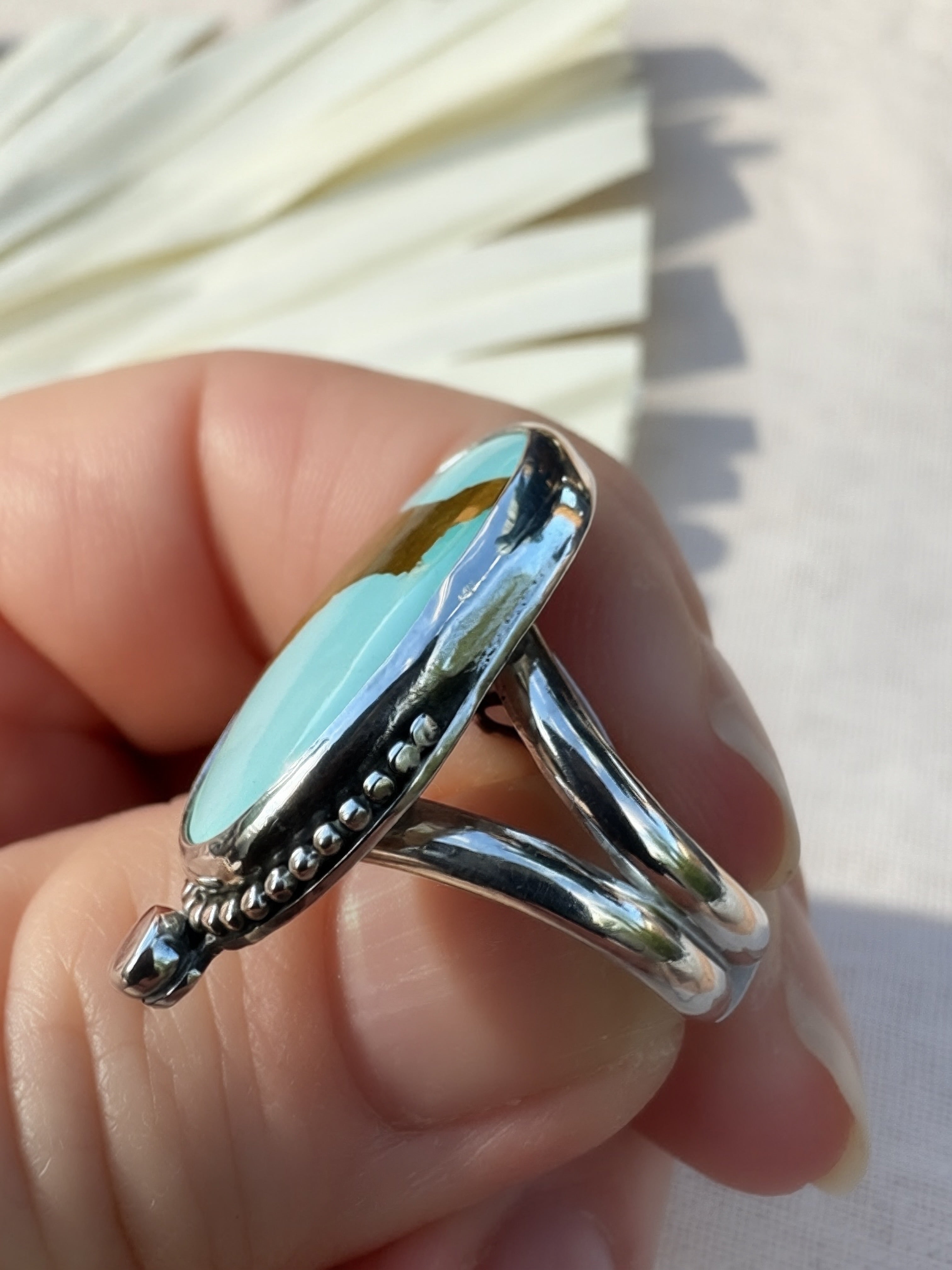 A sterling silver ring with a large, polished oval turquoise light blue stone being held.The ring features a textured beaded border and a split shank band, shown against a neutral light background with a soft-focus fan in the upper left.