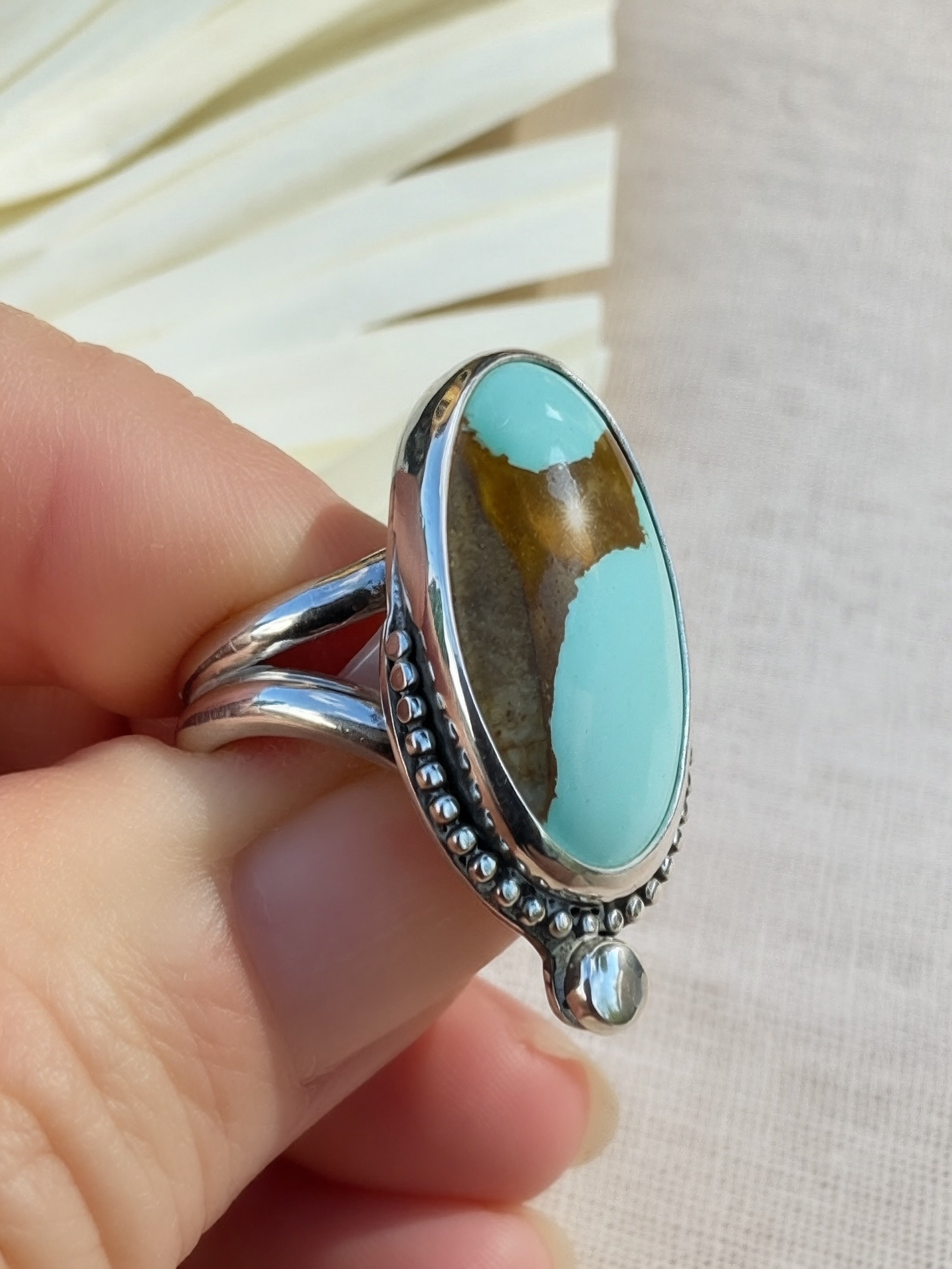 A sterling silver ring with an oval turquoise and brown stone, held on a person's fingers against a light beige fabric background.
