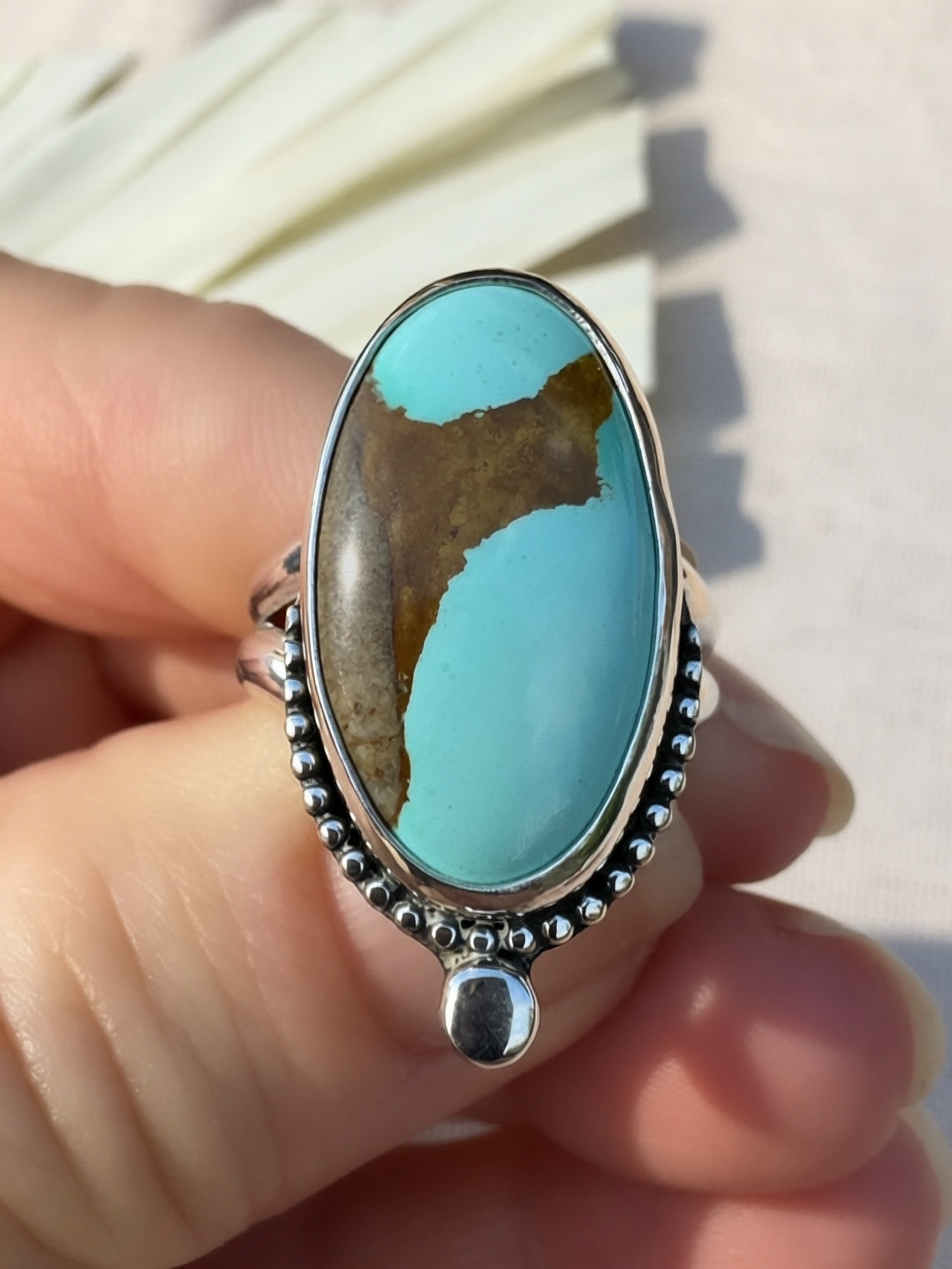 A hand holding a sterling silver ring featuring a large oval turquoise stone with a brown matrix pattern, set against a light background.