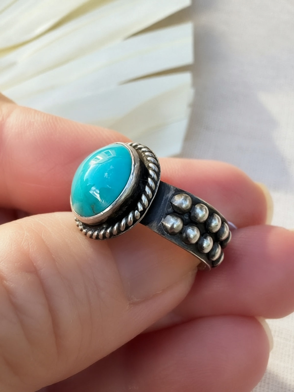A sterling silver ring with a round turquoise cabochon stone, held on a person's fingers. The ring features a textured twisted border around the stone and a band decorated with small silver beads.