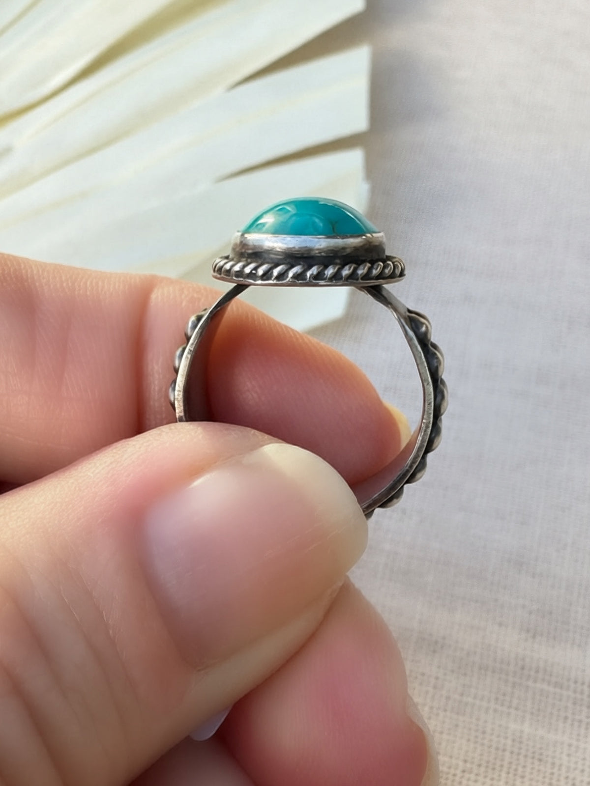 A close-up of a hand holding a silver ring with a round turquoise cabochon stone, set against a soft beige fabric background.