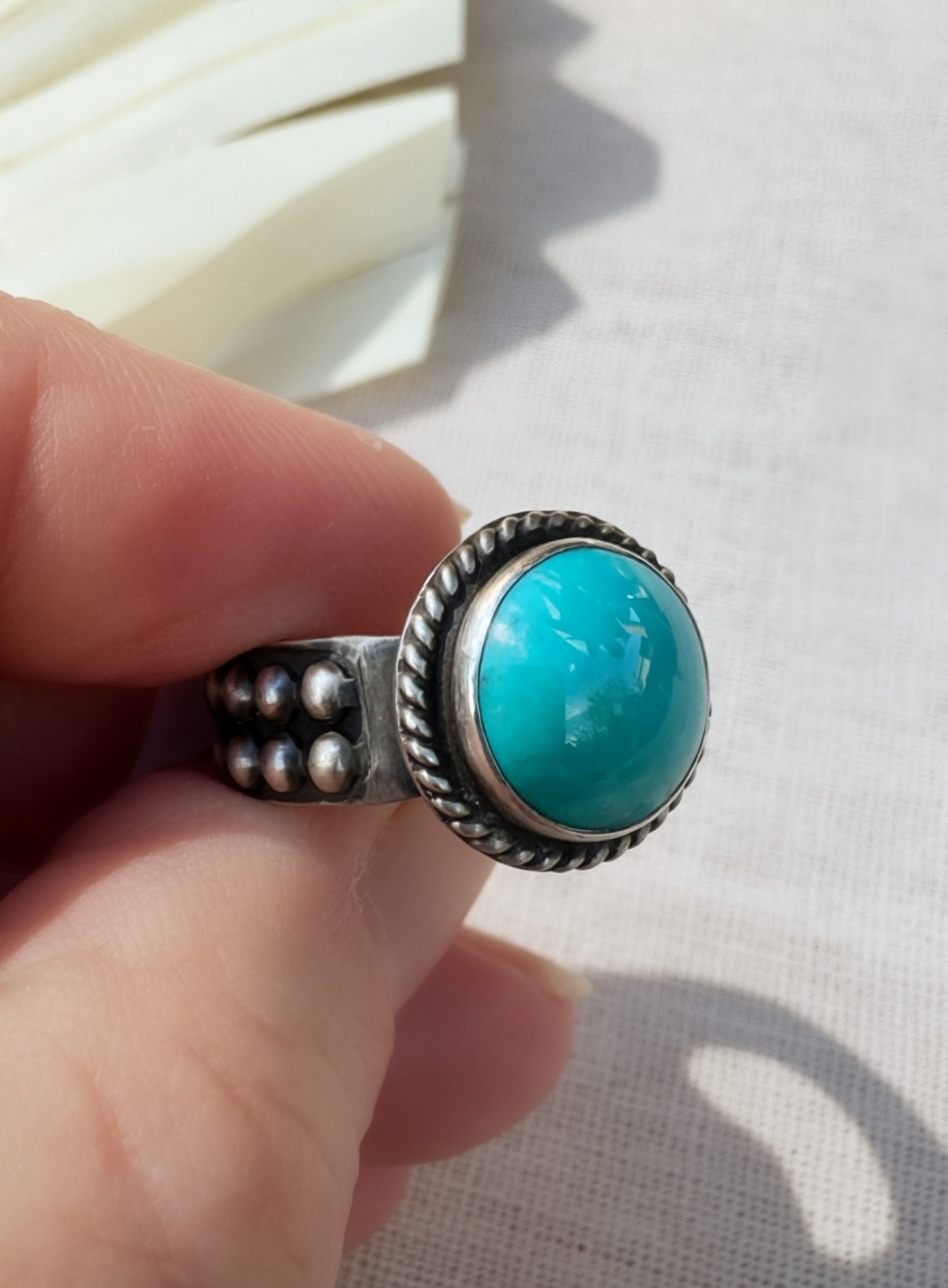 A silver ring with a large round turquoise cabochon gemstone, held between fingers against a light beige textured fabric background.