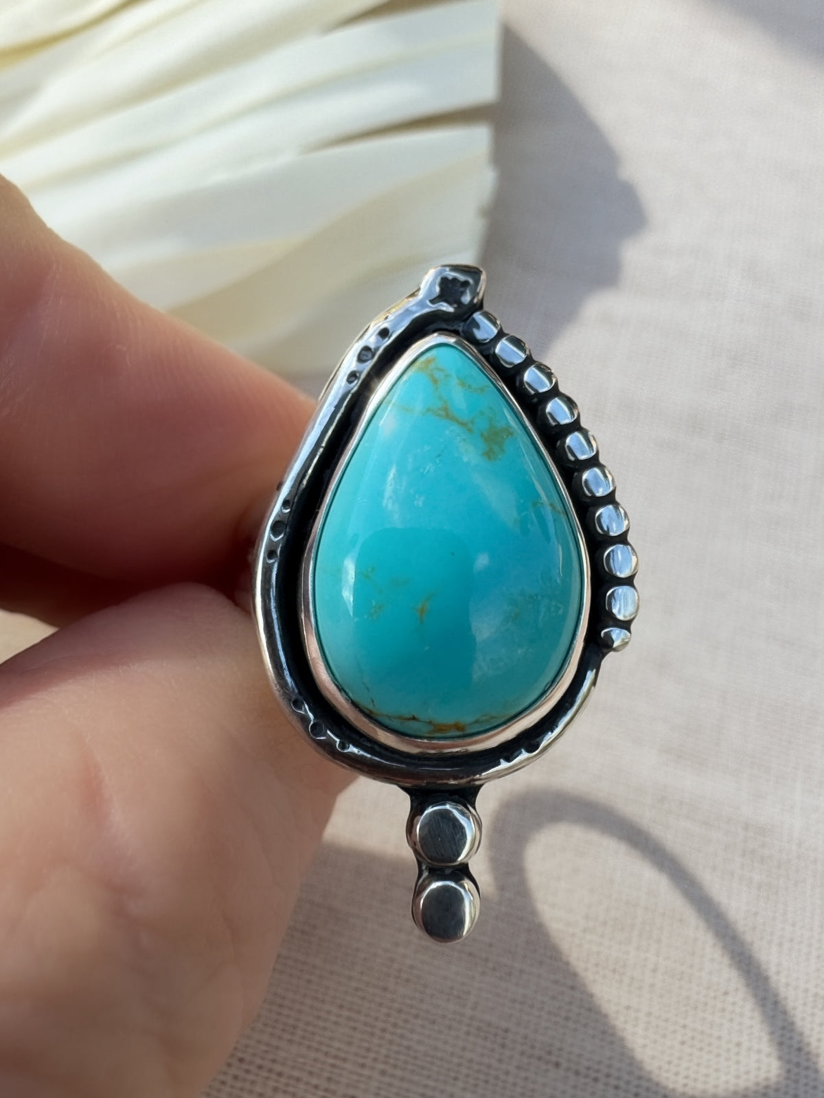 A sterling silver ring with a teardrop-shaped turquoise stone and a snake detail, held between fingers against a neutral beige fabric background.
