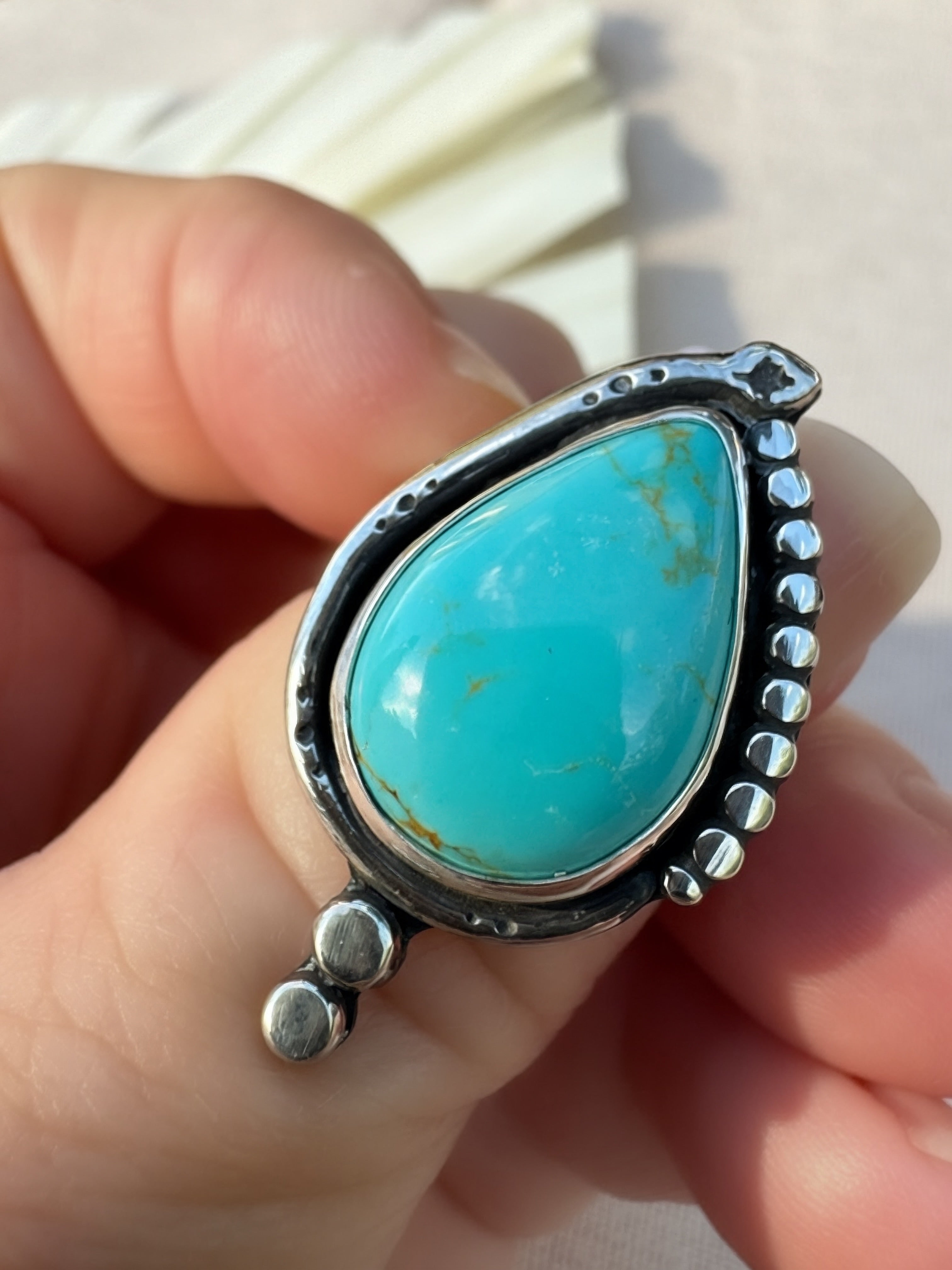 A close-up of a hand holding a sterling silver ring featuring a large, polished turquoise stone with pale natural brown matrix patterns.